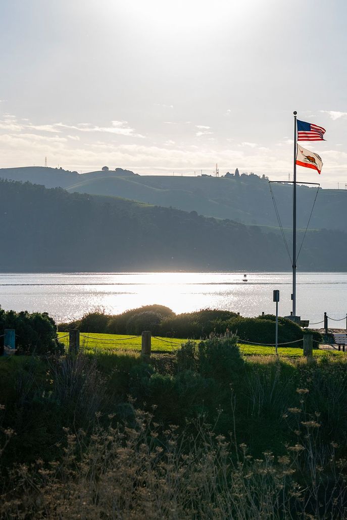 Photo of flagpole at the point of Benicia's Ninth Street park, with Carquinez Strait and hills in the back ground and the sun shining straight into the camera for dramatic effect.  The United States and California flags are on the flagpole