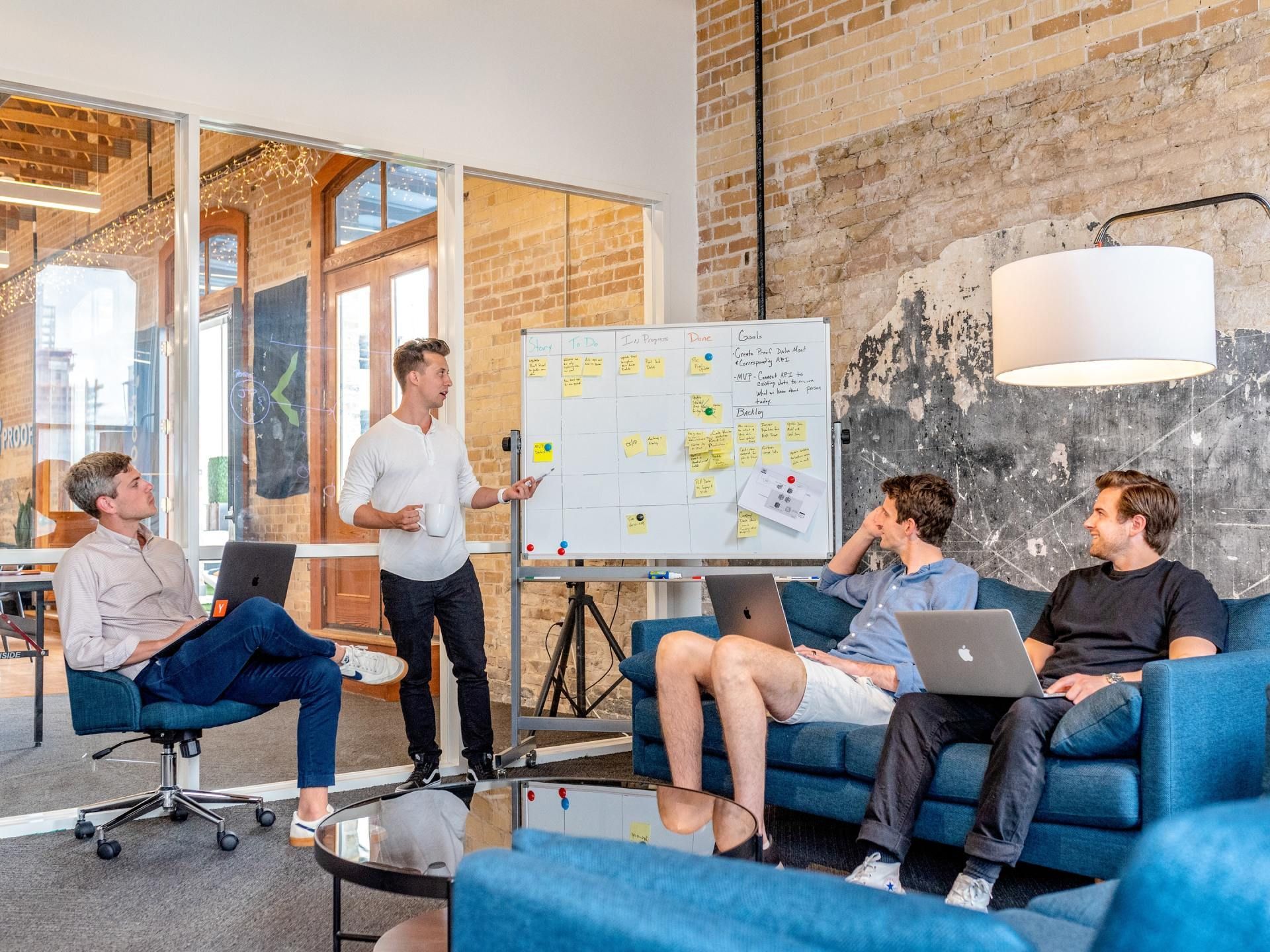 A group of men are sitting on a couch and a man is standing in front of a whiteboard.