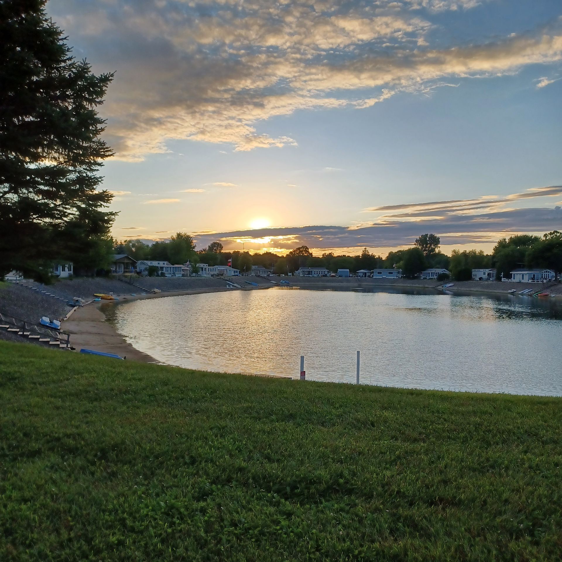A sunset over a calm lake with a grassy foreground, a sandy shore, and trees and houses in the distance.