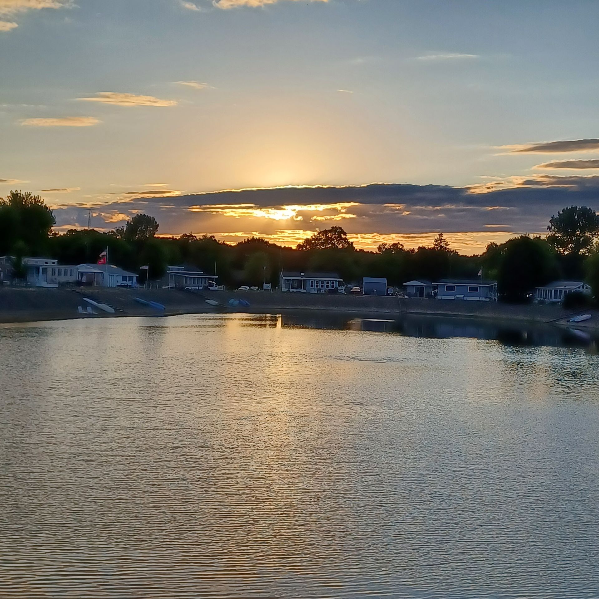 A sunset casts a golden glow over a calm lake, with silhouettes of houses and trees along the distant shoreline.