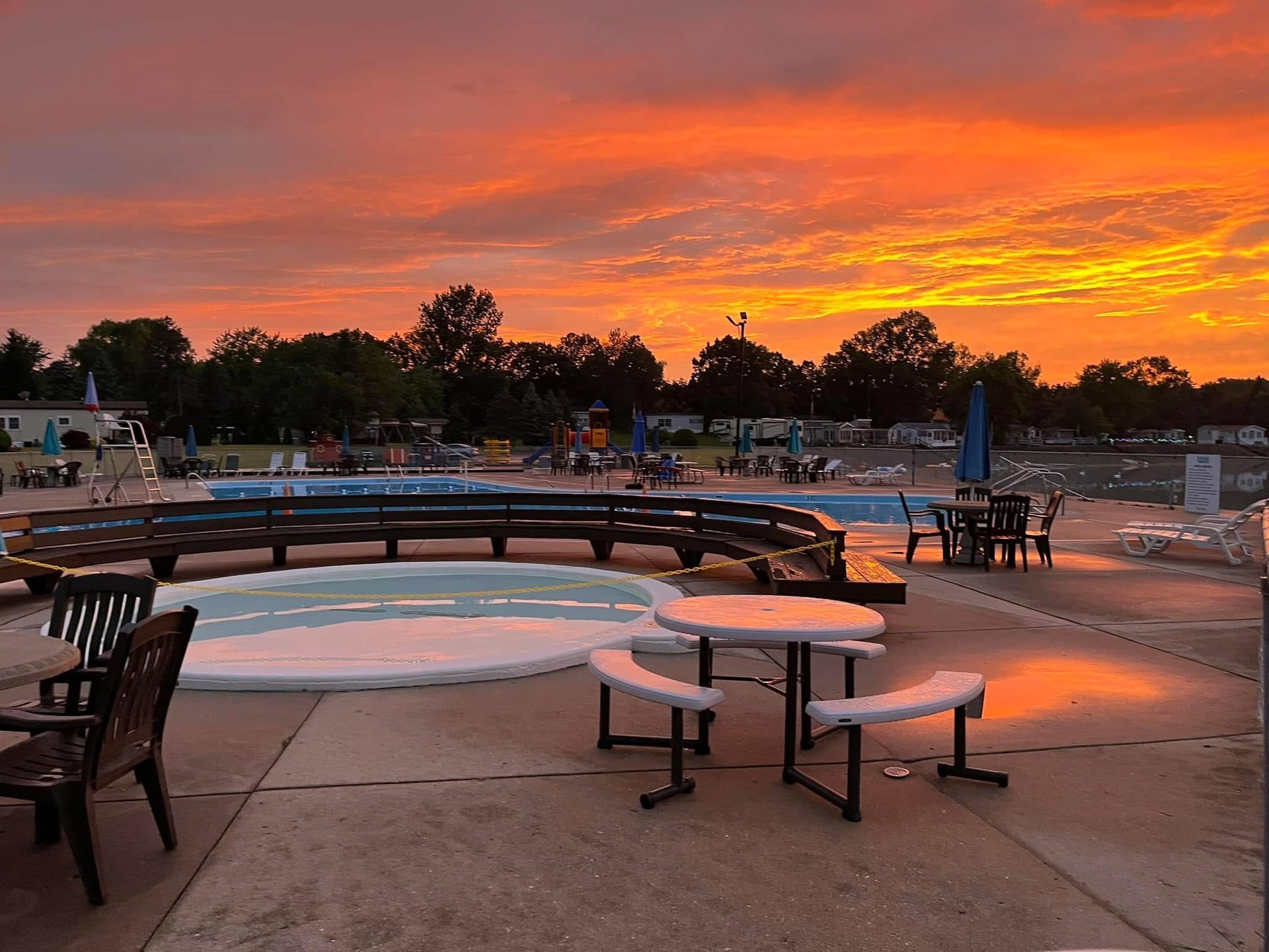 Outdoor patio with tables and a pool at sunset, featuring a vibrant orange and purple sky over trees and deck furniture.