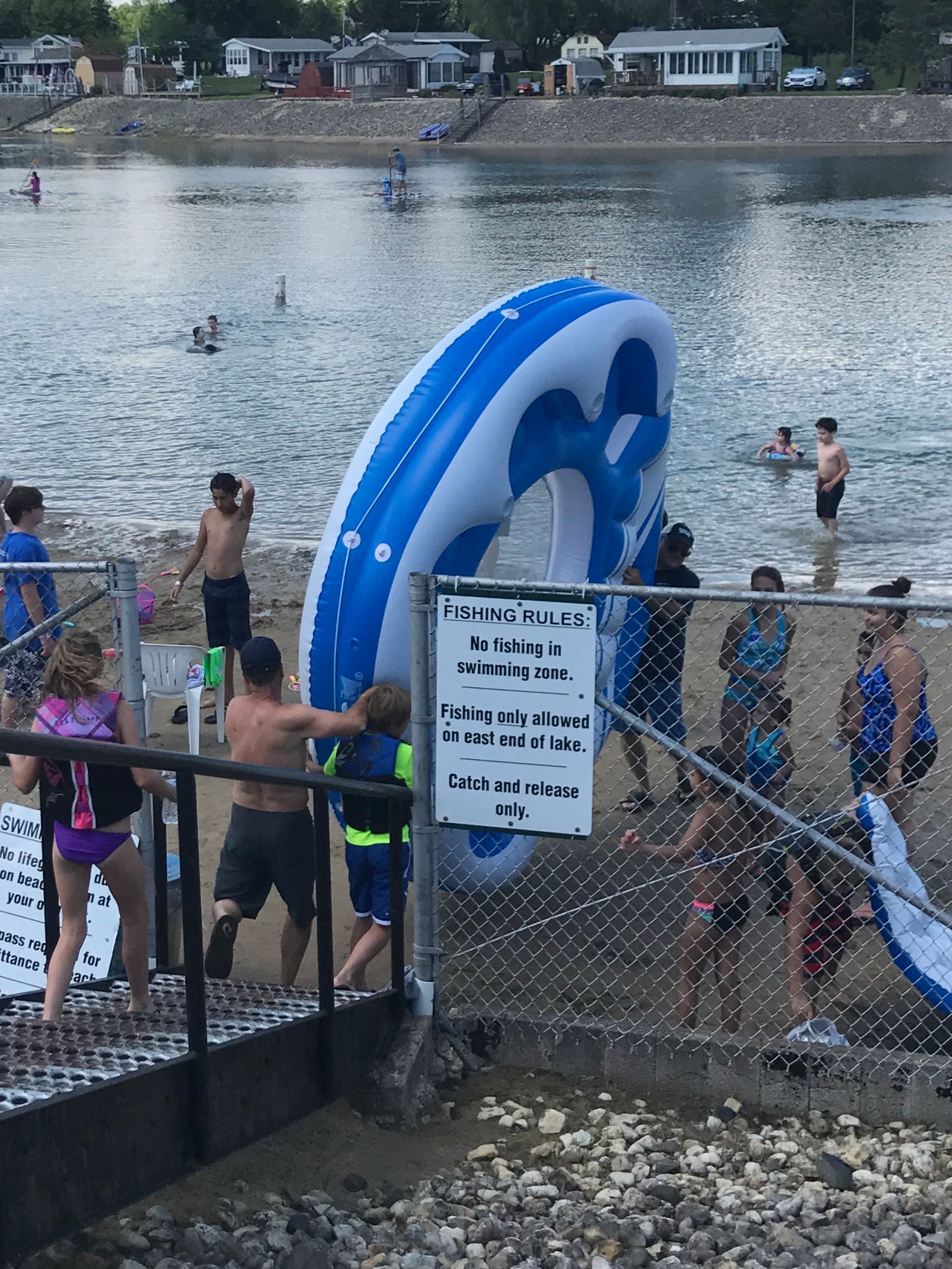 People carry a large, blue-and-white inflatable ring down stairs toward a beach where others swim in the water.