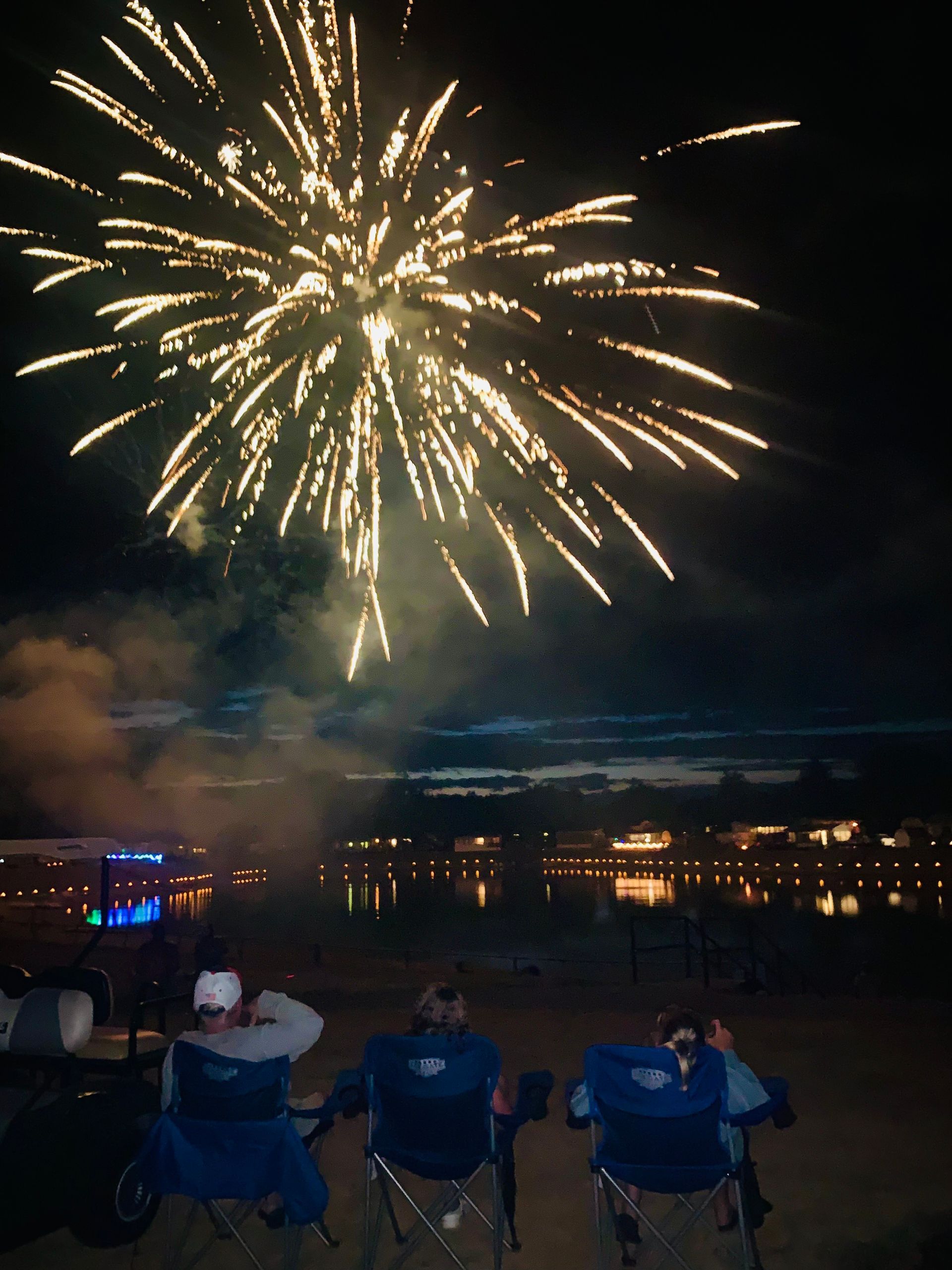 Three people sit in folding chairs watching a bright gold firework explode over a lake at night.