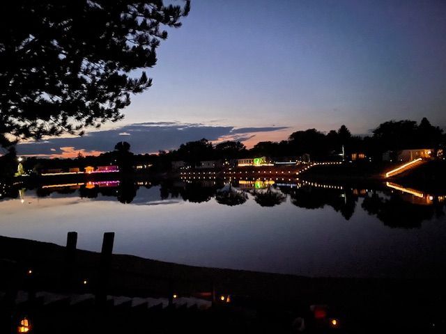 A tranquil lake at dusk with trees and illuminated buildings reflected in the calm water under a sunset sky.