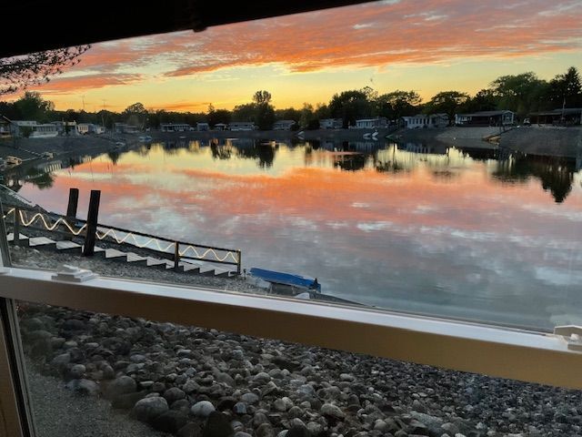 A vibrant, orange sunset reflects on calm lake water, viewed from a window overlooking a rocky shoreline and a small dock.