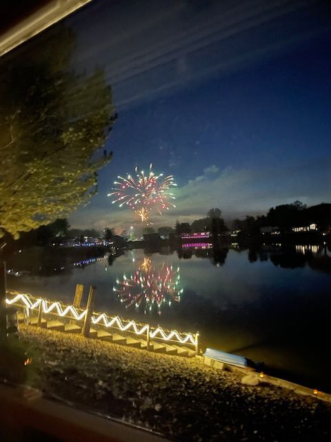 Fireworks explode over a calm lake at twilight, their vibrant colors reflecting on the water near a lit shoreline.