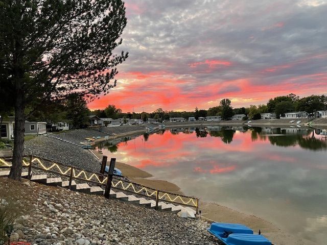 A vibrant red and orange sunset reflects off a calm pond, bordered by a rocky shoreline and a line of mobile homes.