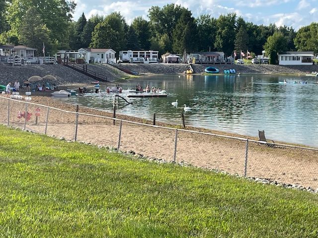 A sandy beach with a swimming area, floating platforms, and small homes on a hill overlooking the water.