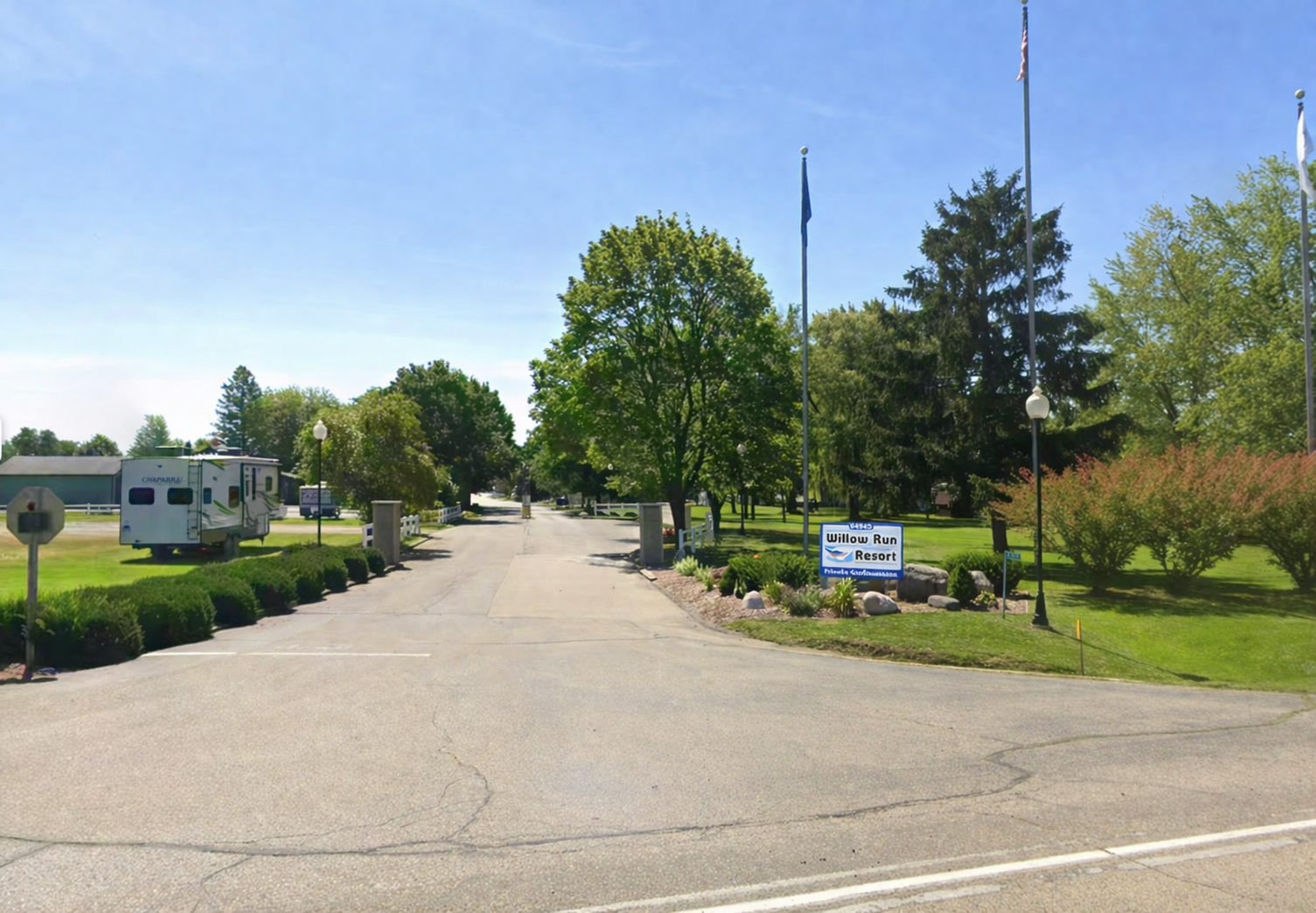 A paved entrance to a park or campground with mature green trees, a camper trailer, and flags under a bright blue sky.