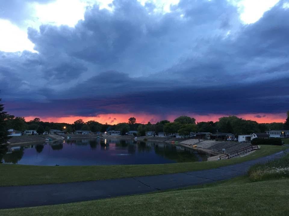 A vibrant sunset glows bright orange behind dark storm clouds over a calm pond in a residential area.