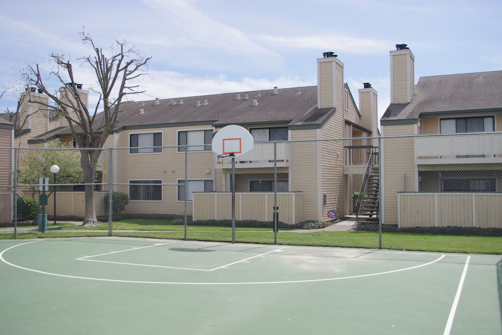 Outdoor basketball court with hoop in front of apartment buildings and a leafless tree