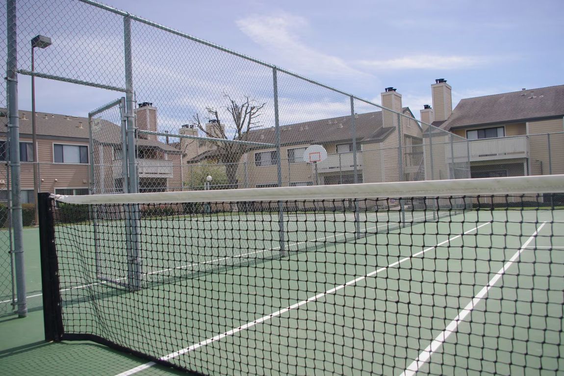 Empty tennis court with net, chain-link fence, and apartment buildings in the background