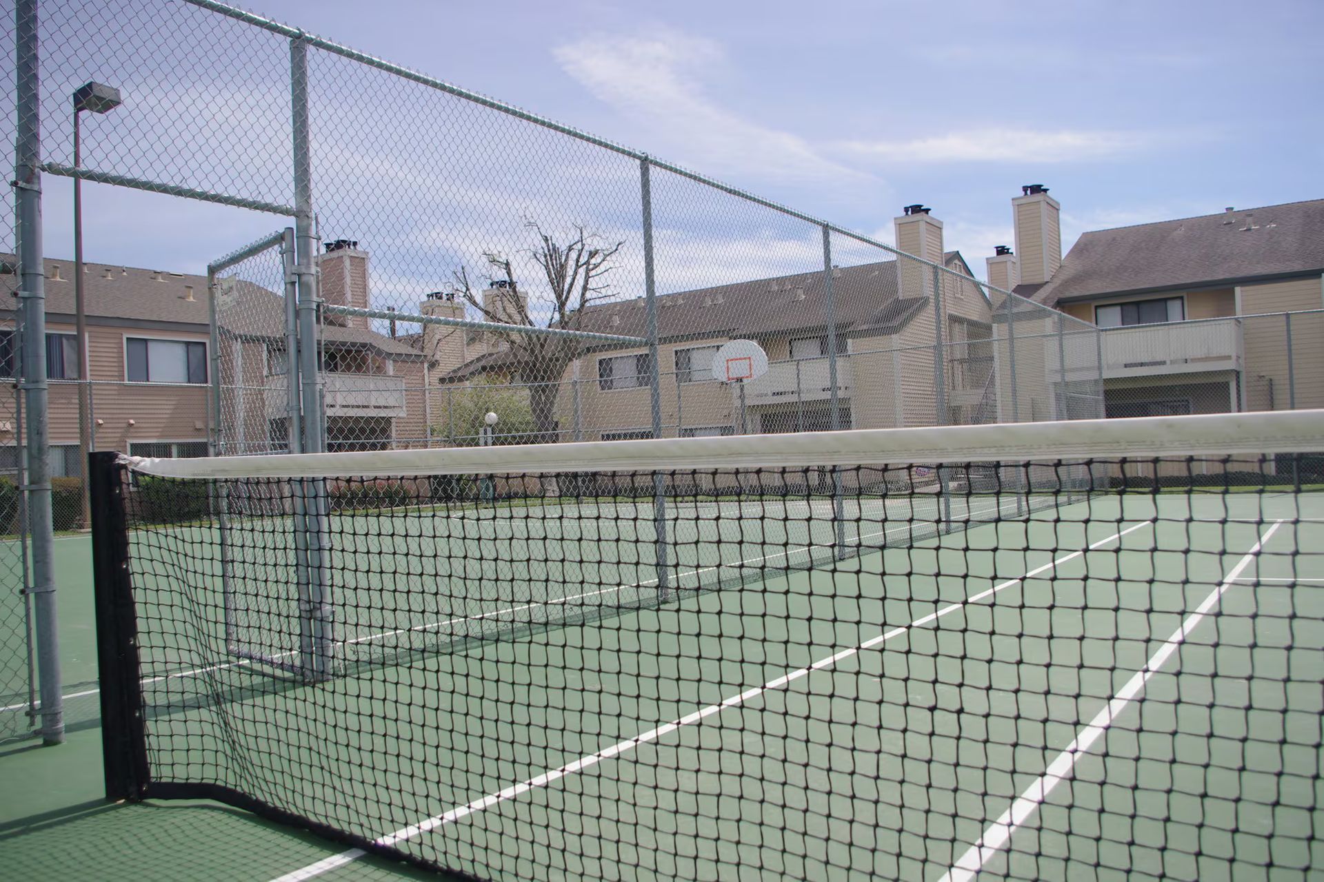 Empty tennis court with net, surrounded by a chain-link fence and apartment buildings.