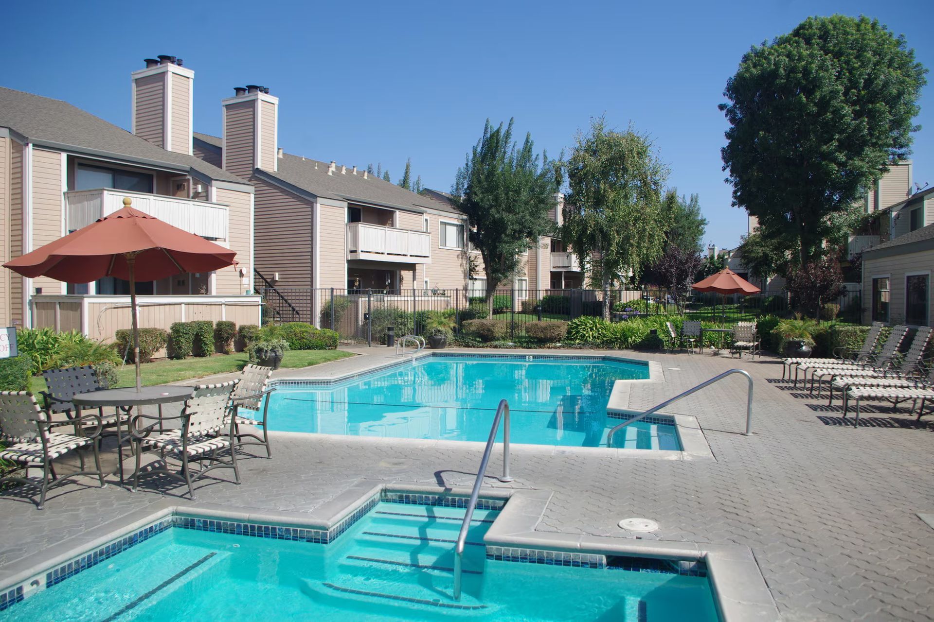 Apartment pool courtyard with blue swimming pools, lounge chairs, and red umbrellas under a clear sky