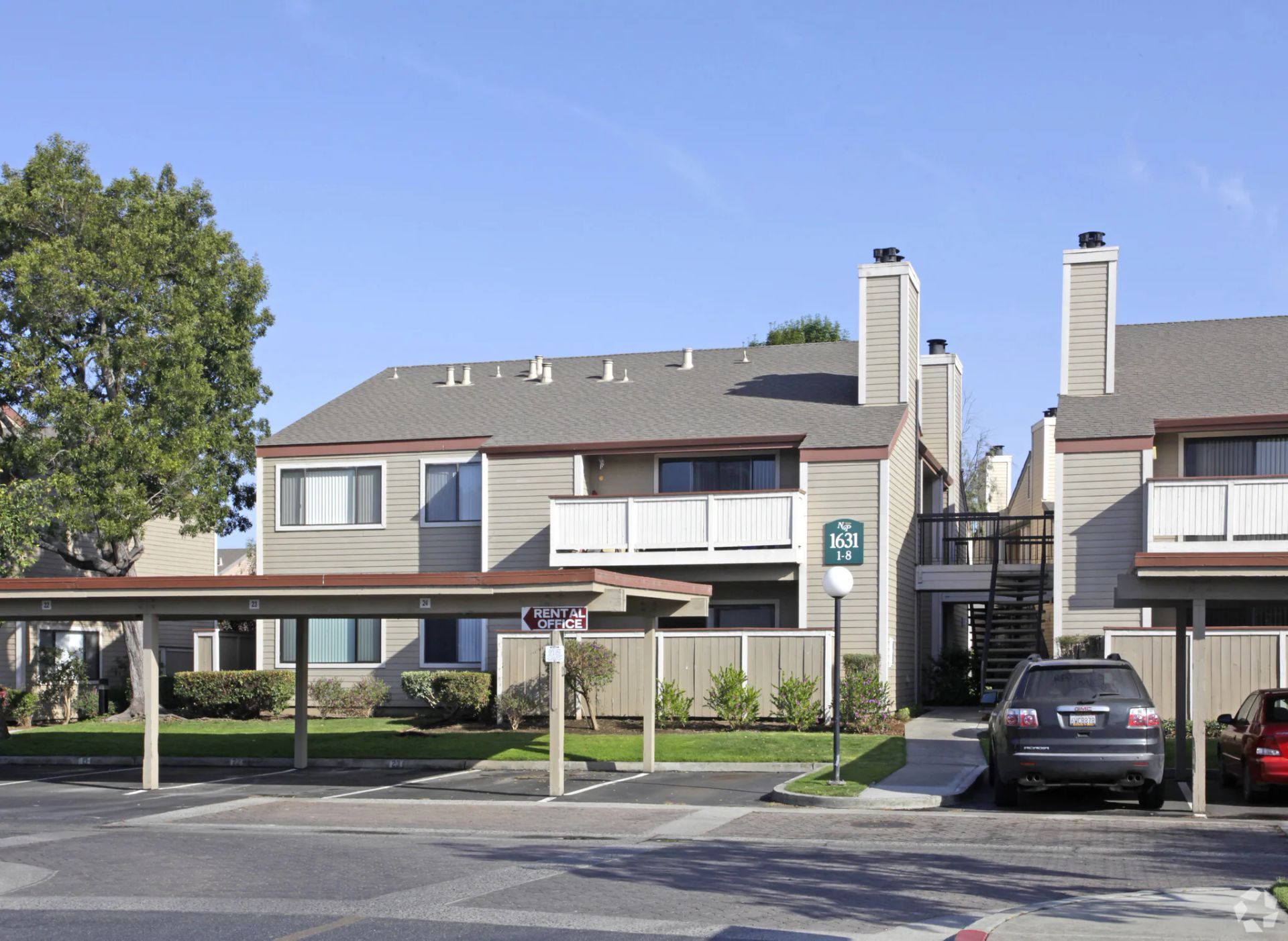 Apartment complex with covered parking, balconies, stairs, and a parked black car under a blue sky