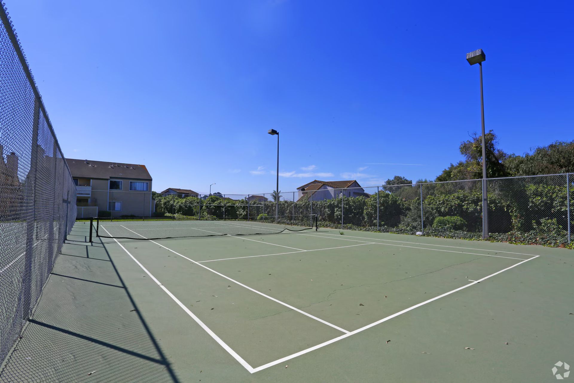 Empty outdoor tennis court with green surface, chain-link fence, and clear blue sky
