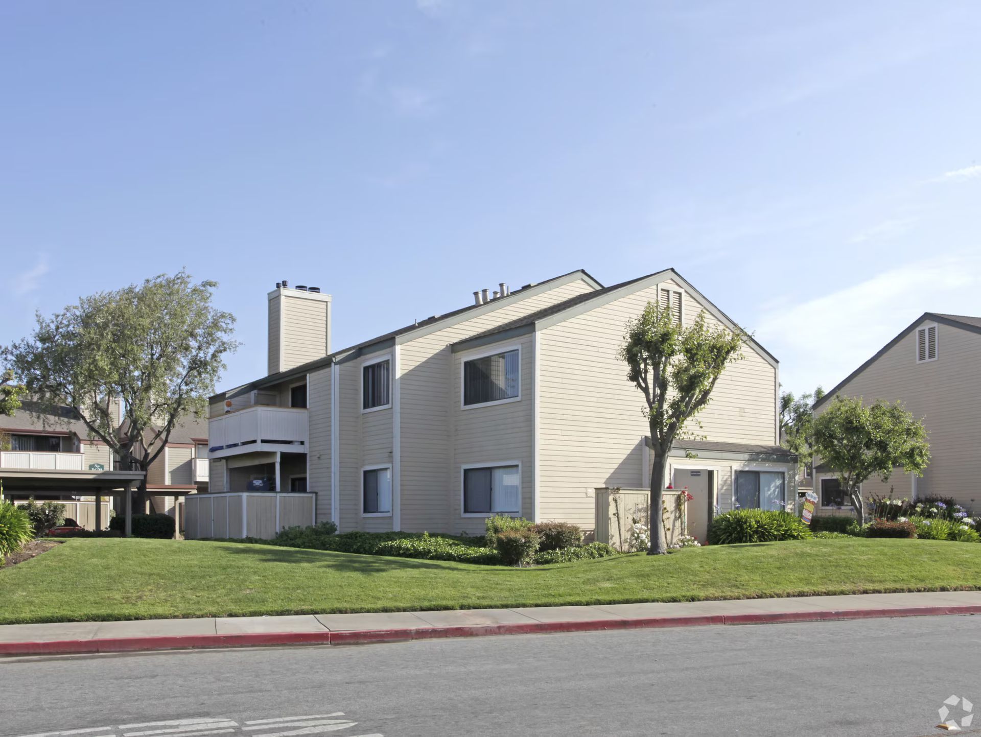 Residential apartment building with beige siding, green lawn, and trees along a quiet street.