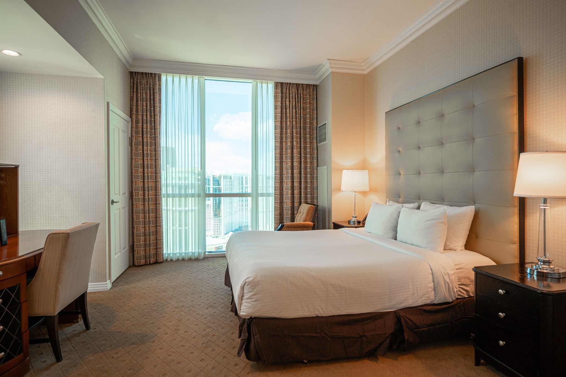 Hotel room with a king bed, desk, lamps, and balcony view through tall curtains