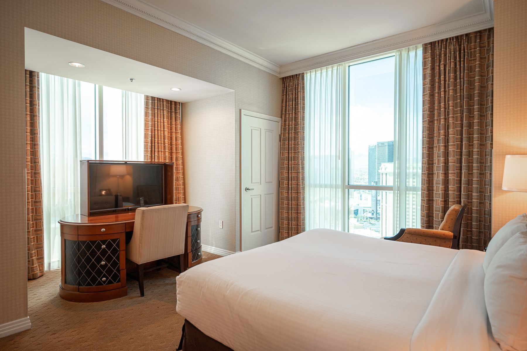 Bright hotel room with twin beds, TV, desk, and large windows with sheer curtains.