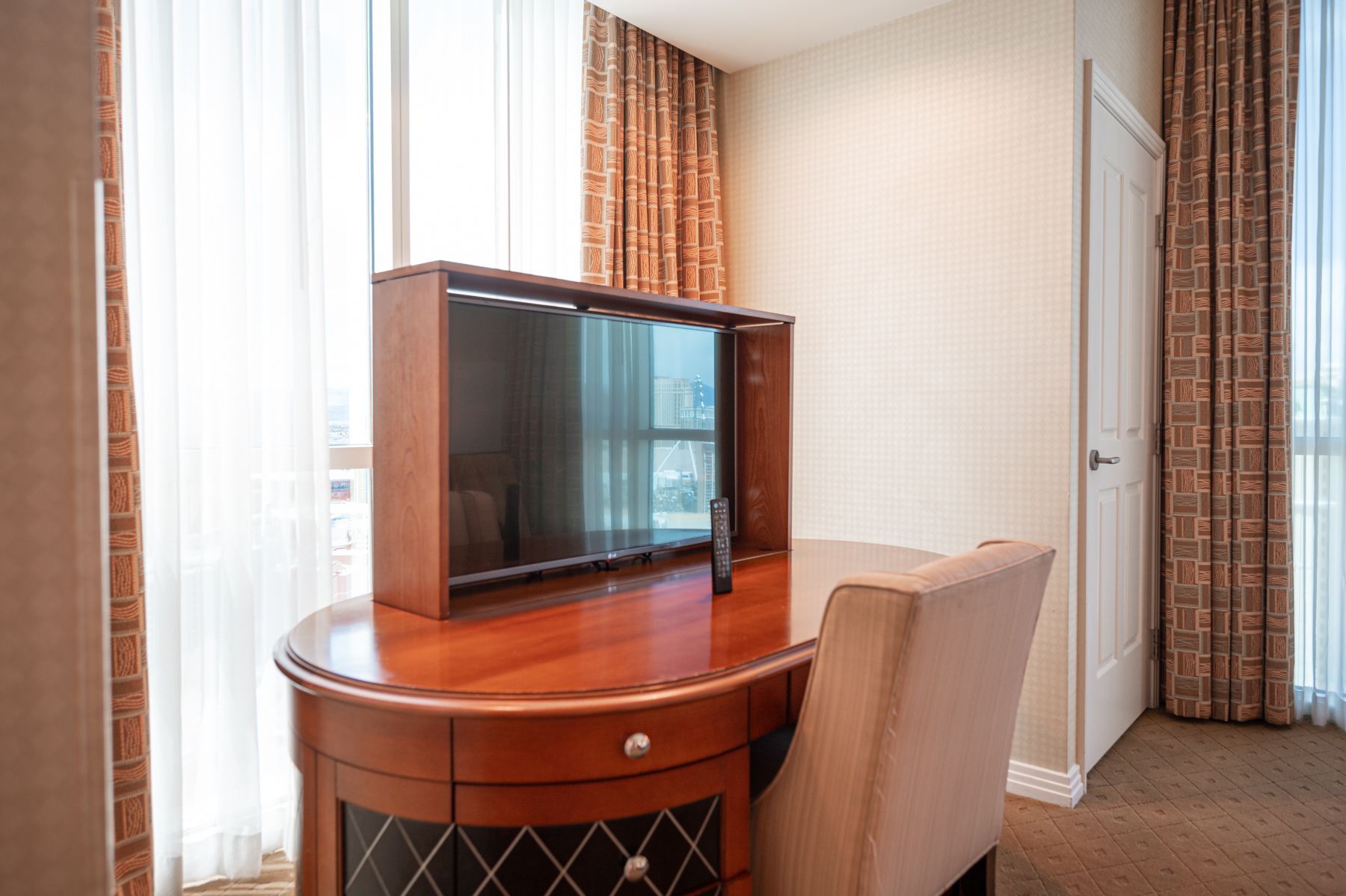 Hotel room corner with TV on a wooden table beside a chair and curtains.
