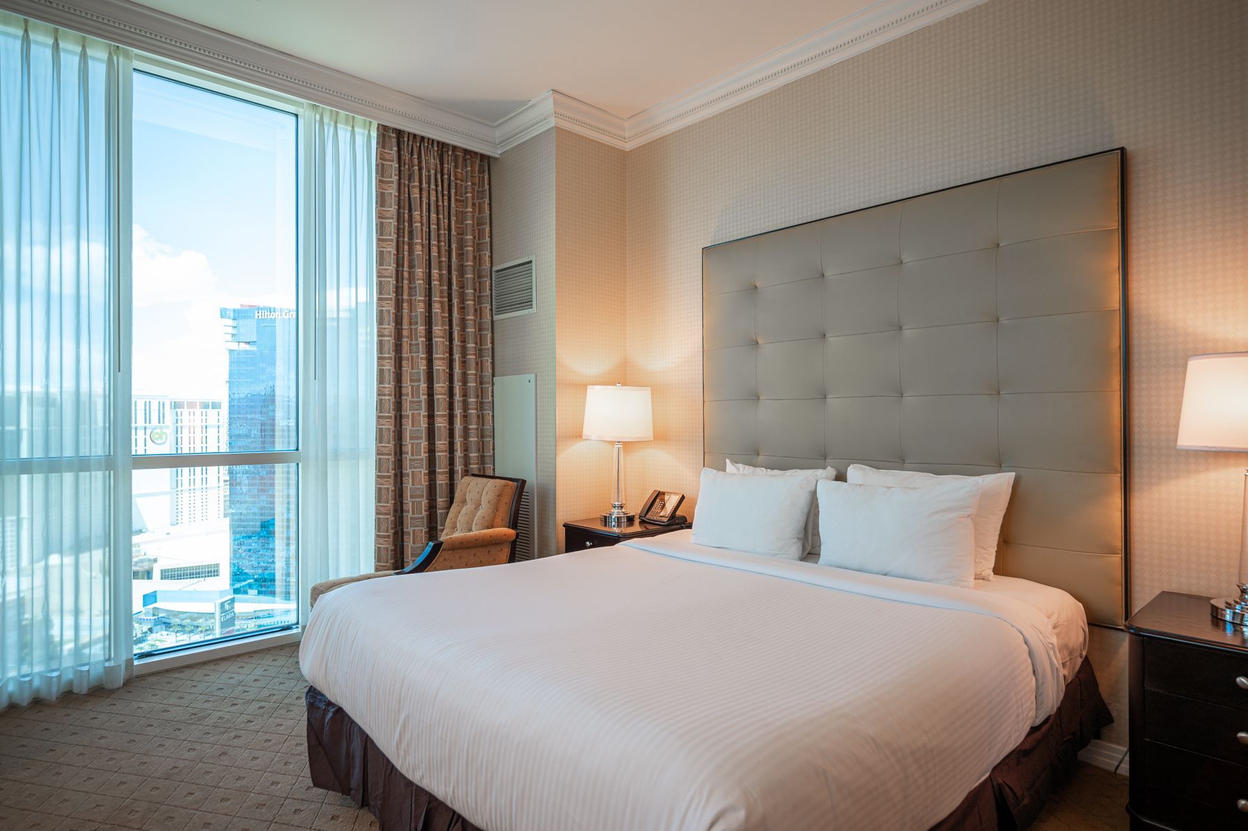 Modern hotel room with king bed, tufted headboard, lamp, chair, and floor-to-ceiling windows overlooking a city view