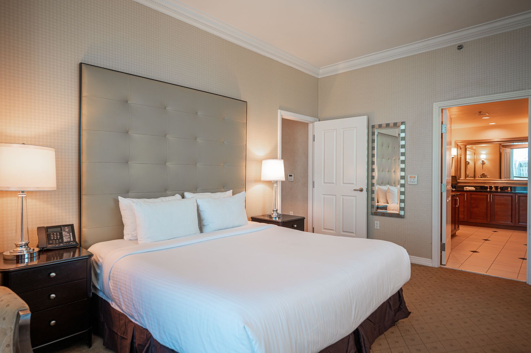 Bright bedroom with a large bed, gray tufted headboard, bedside lamps, and an open doorway to a bathroom.