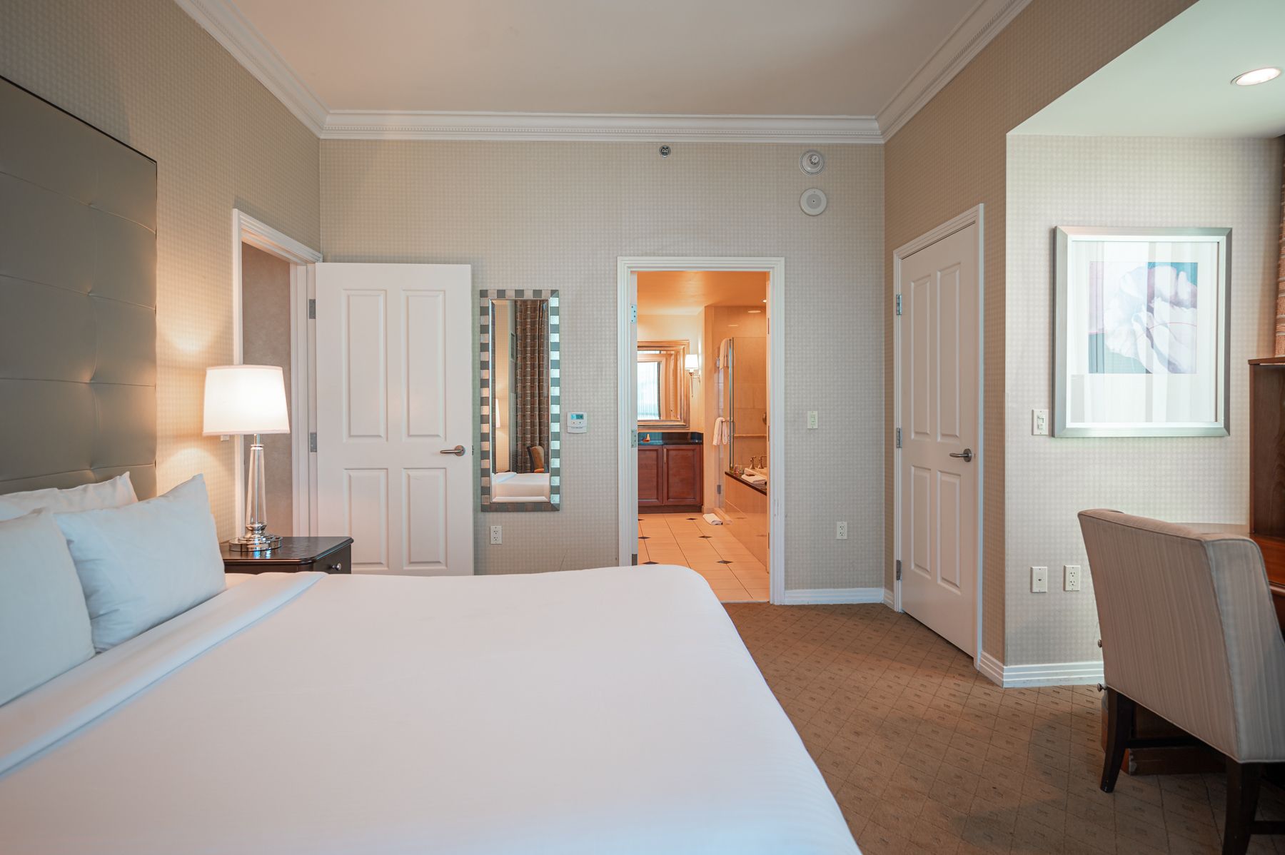 Bright bedroom with a neatly made bed, lamp, chair, and open doorway to a warmly lit bathroom.