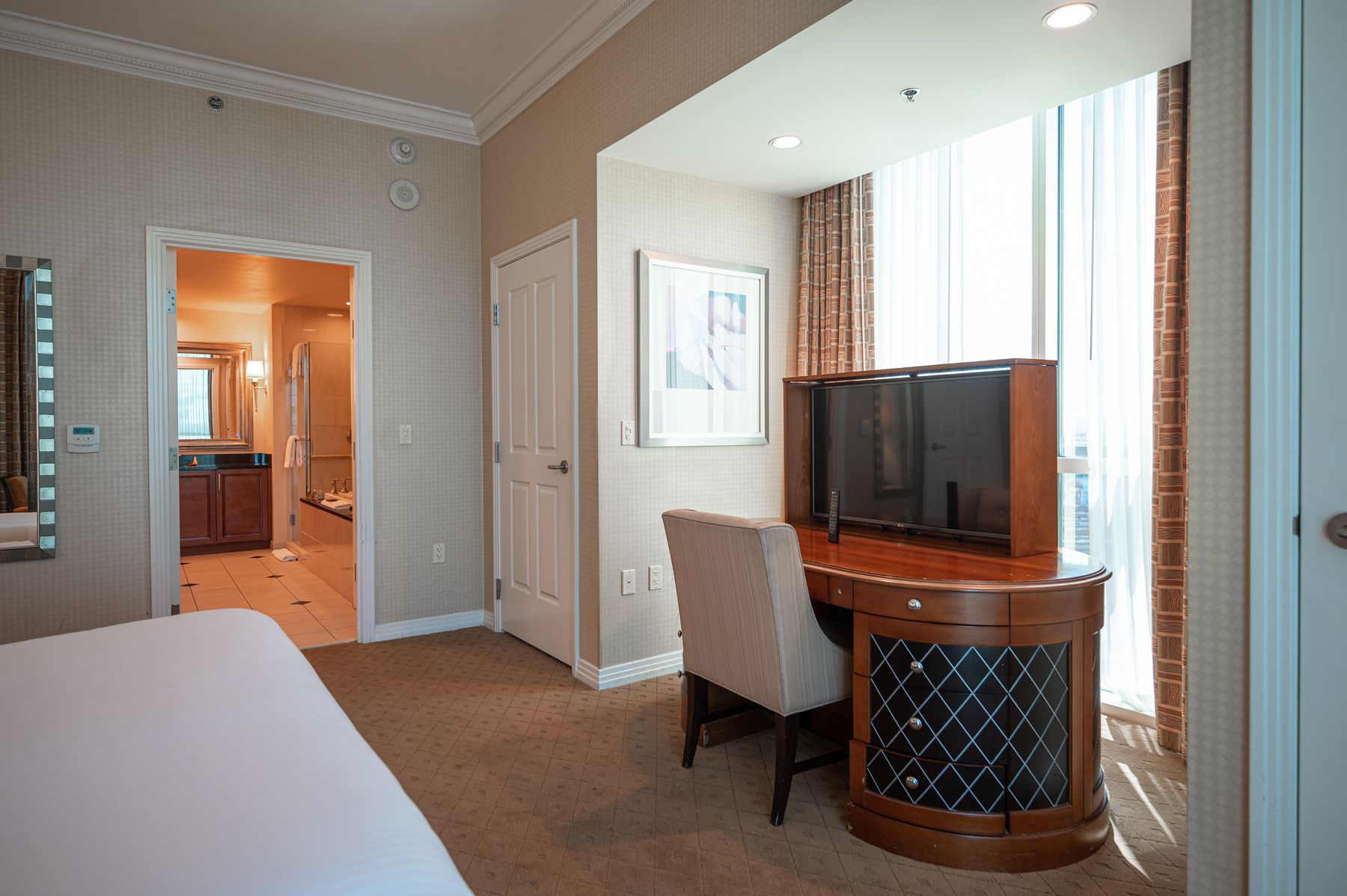 Hotel room with a TV on a corner cabinet, chair, and open doorway to a bathroom and hallway.