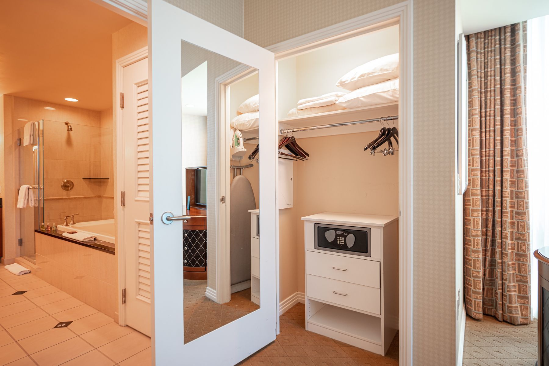 Open closet with shelves, hanging clothes, and a small safe in a bright hotel room hallway.