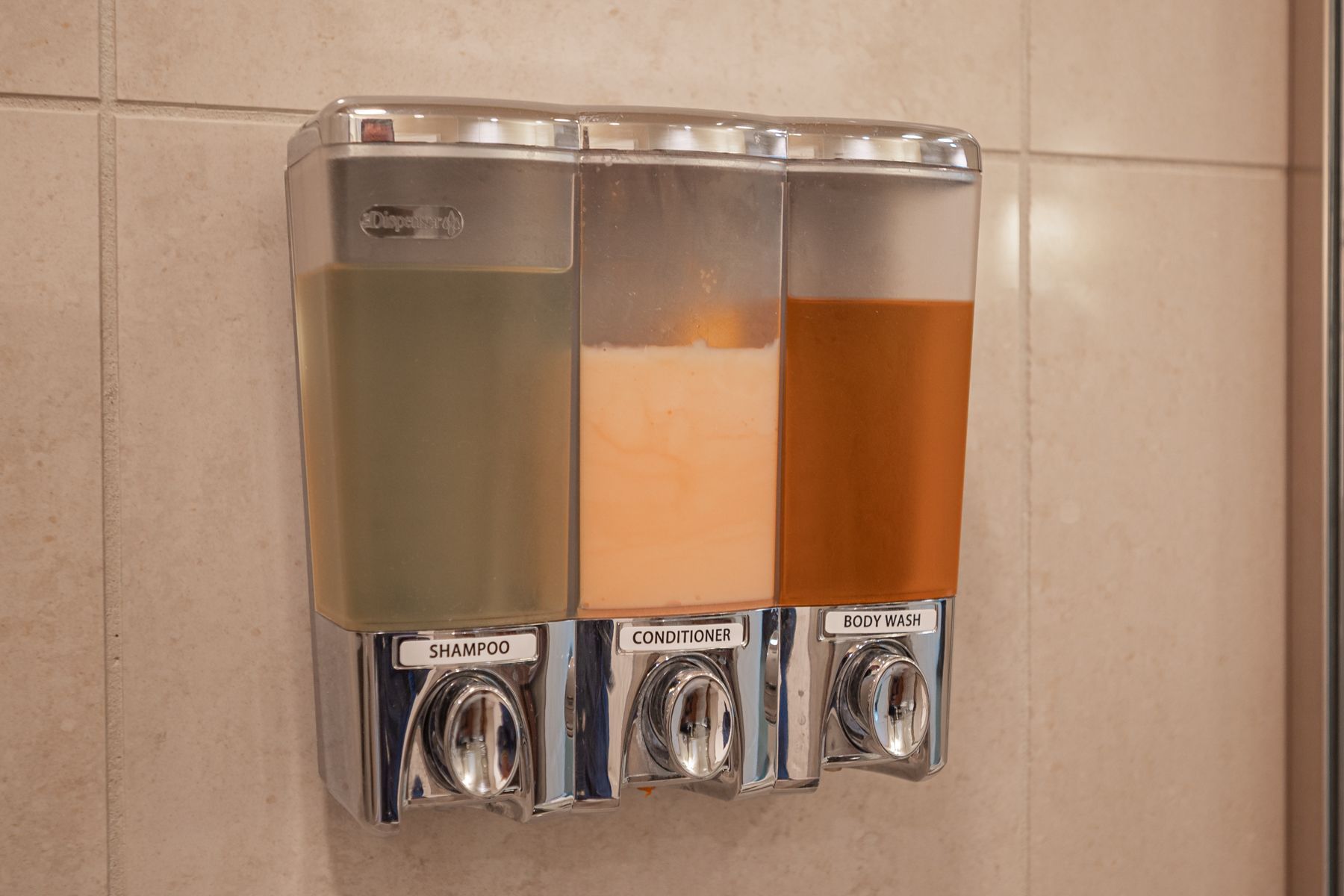 Three wall-mounted soap dispensers in a tiled restroom, filled with green, white, and orange soap.