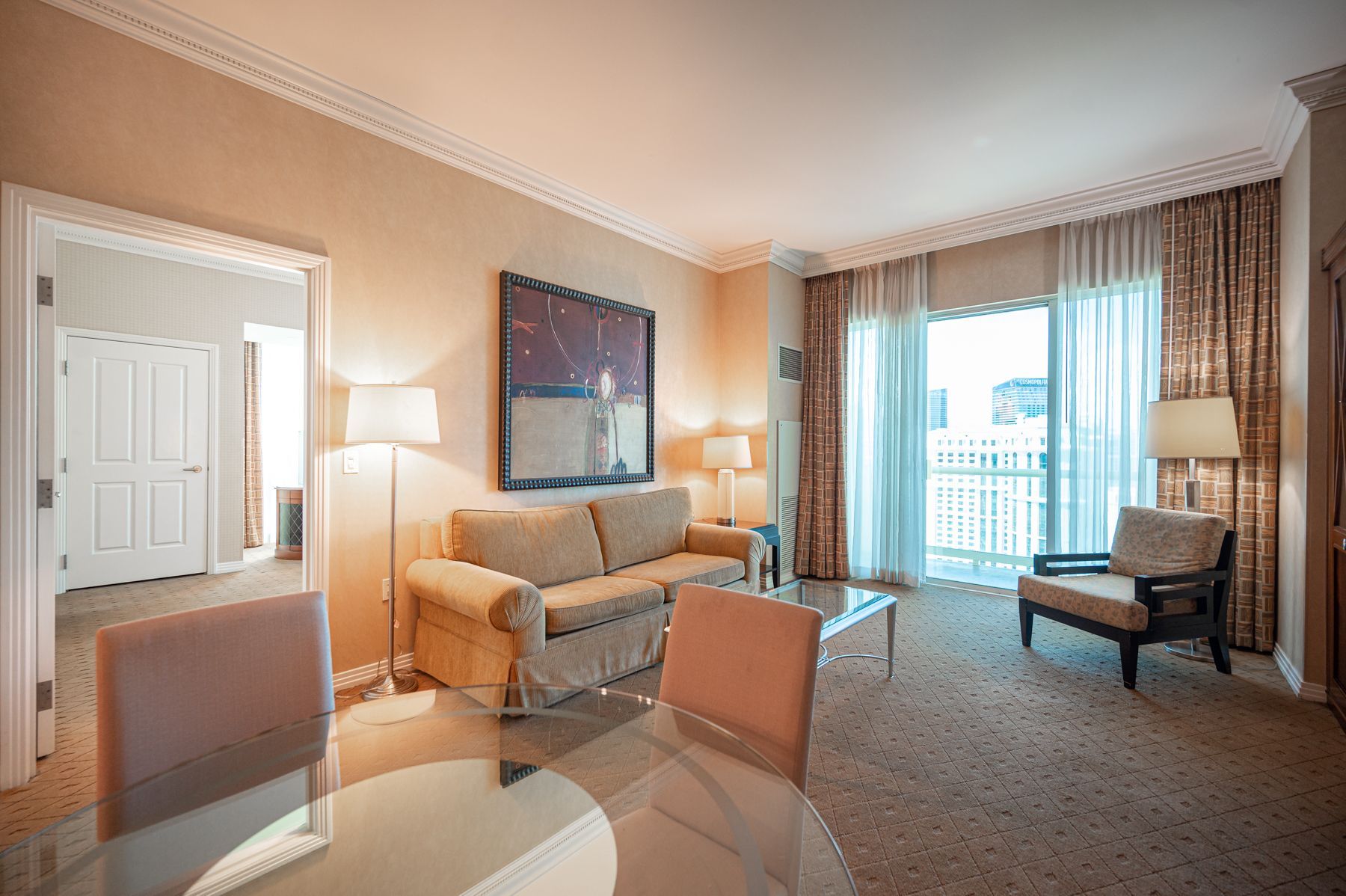 Modern hotel room with sofa, chairs, lamps, and balcony doors overlooking a city view