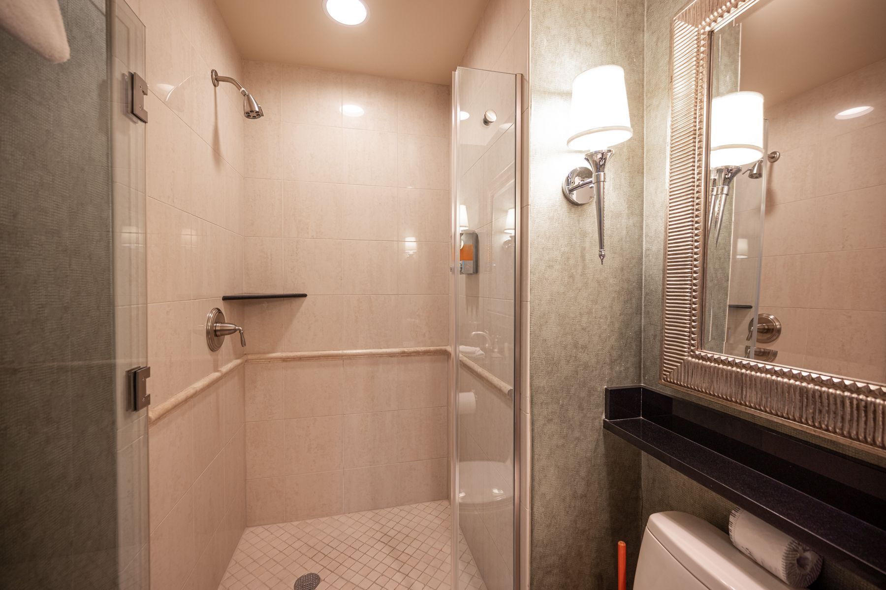 Compact beige bathroom with shower stall, mirror, vanity, and wall sconces