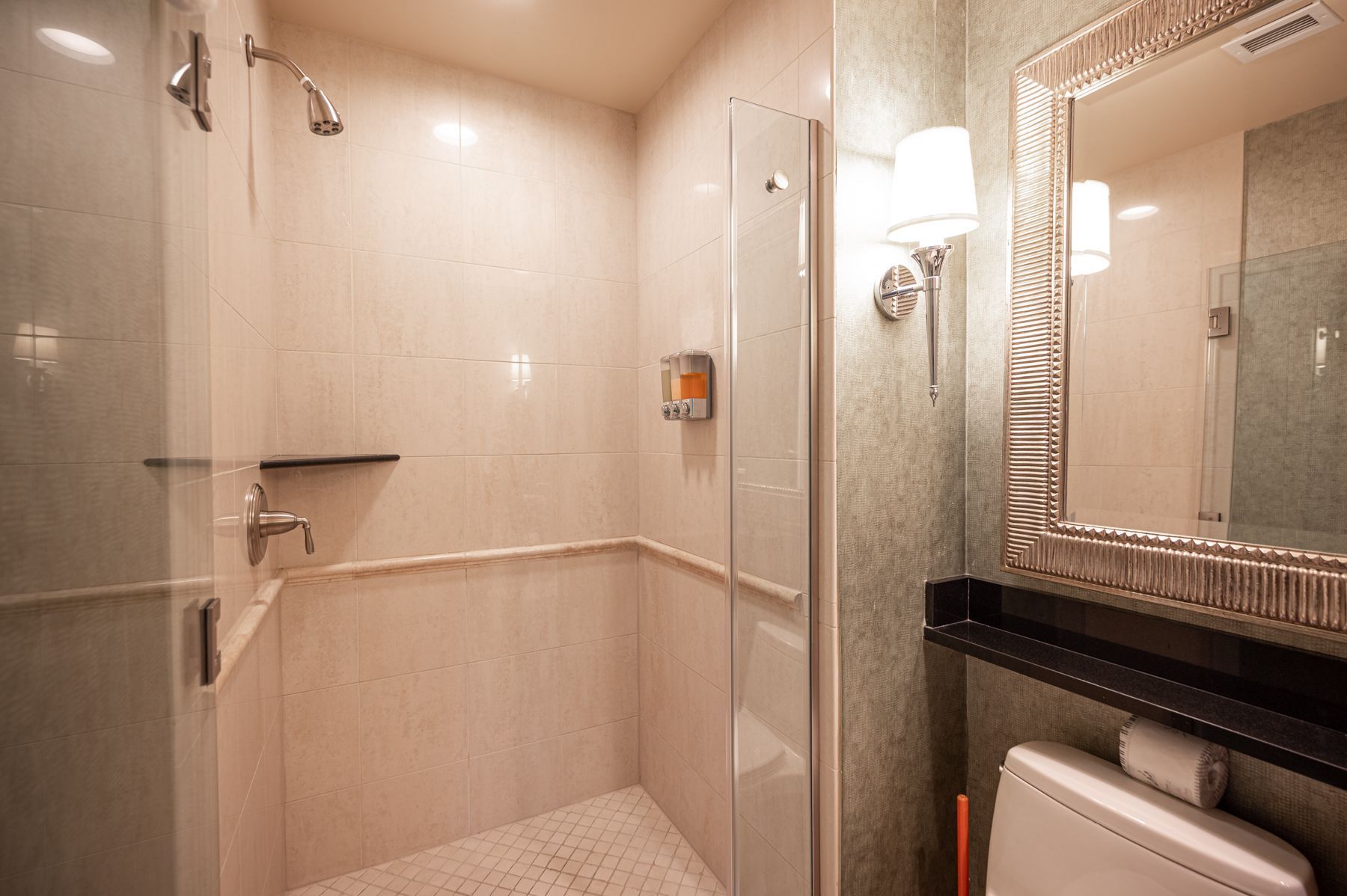Accessible shower and toilet area with beige tile, glass shower doors, and a wall sconce mirror.