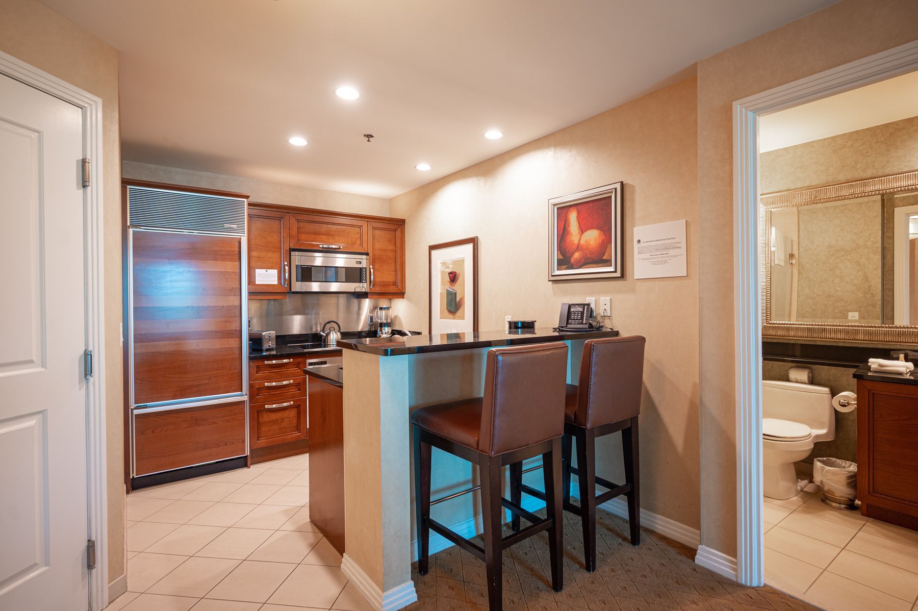 Modern kitchen with bar stools, wood cabinets, and a tiled hallway view into a bathroom