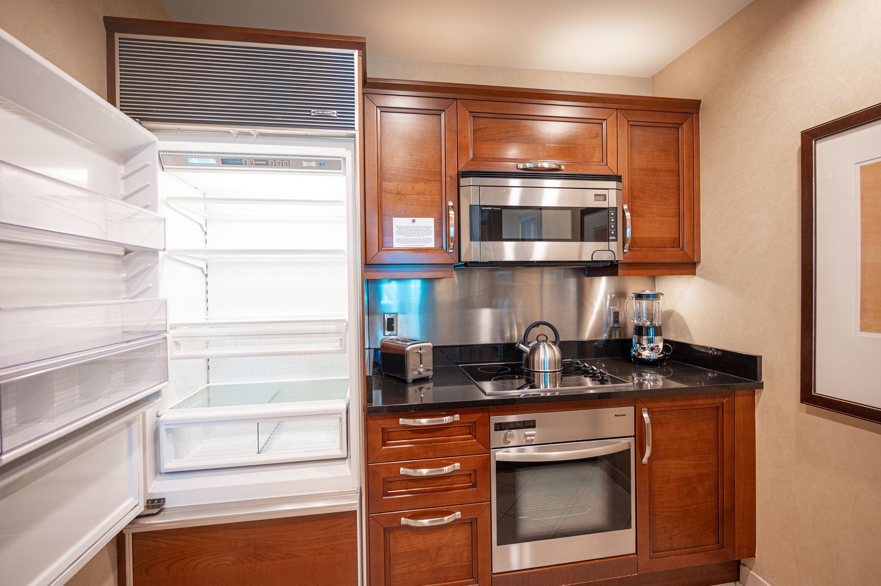 Empty kitchen with open refrigerator, wood cabinets, black countertops, and stainless steel appliances.