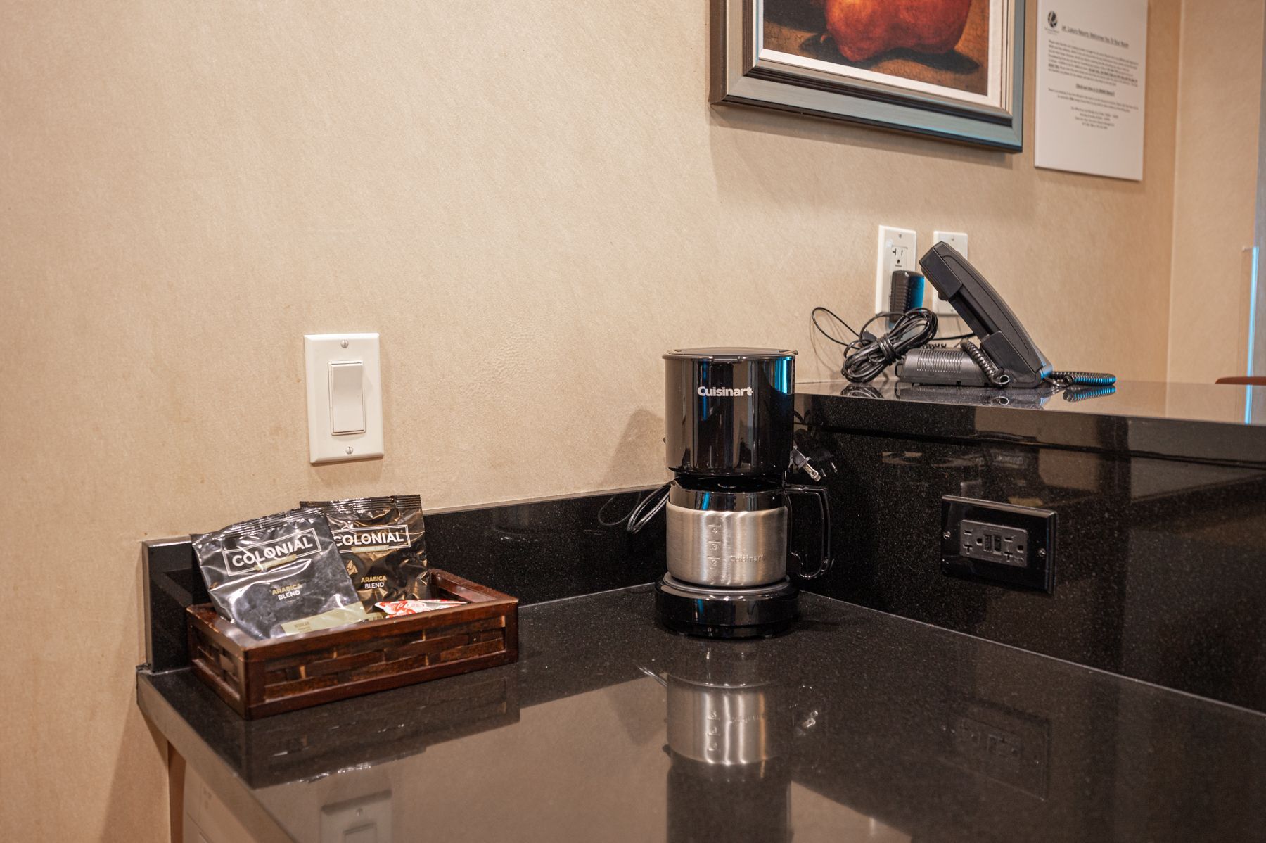 Coffee station with a countertop brewer, black counter, and tray of packaged snacks against a beige wall.