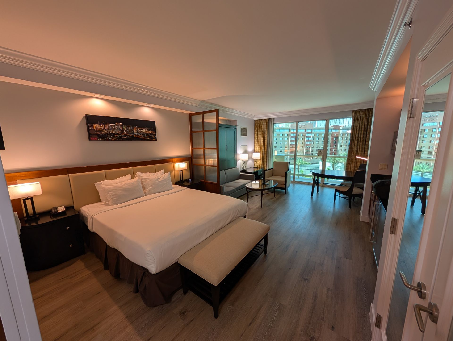 Modern hotel bedroom with a large bed, warm lighting, wood floors, and a balcony view through glass doors