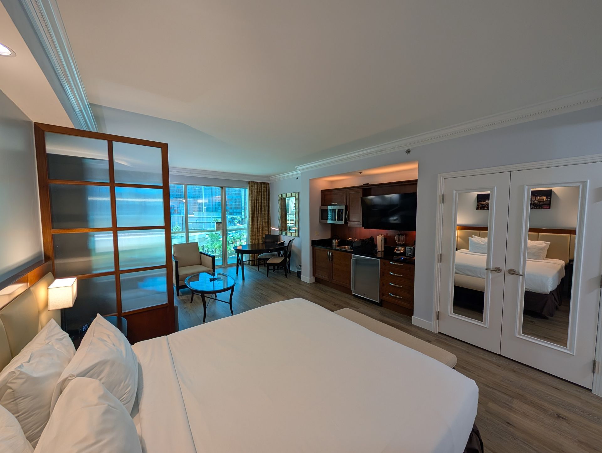 Modern hotel room with king bed, TV, glass wardrobe, desk, and ocean-view windows.