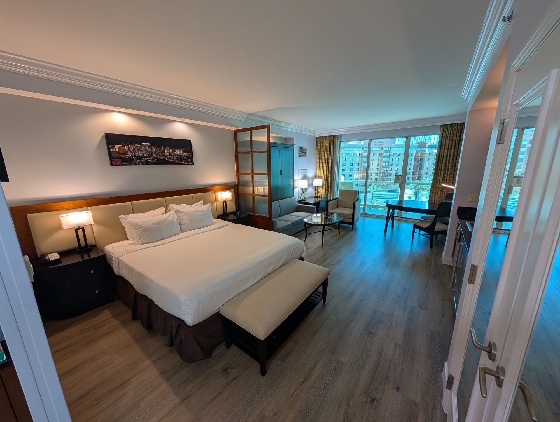 Modern hotel bedroom with king bed, warm lighting, and balcony doors overlooking a city view