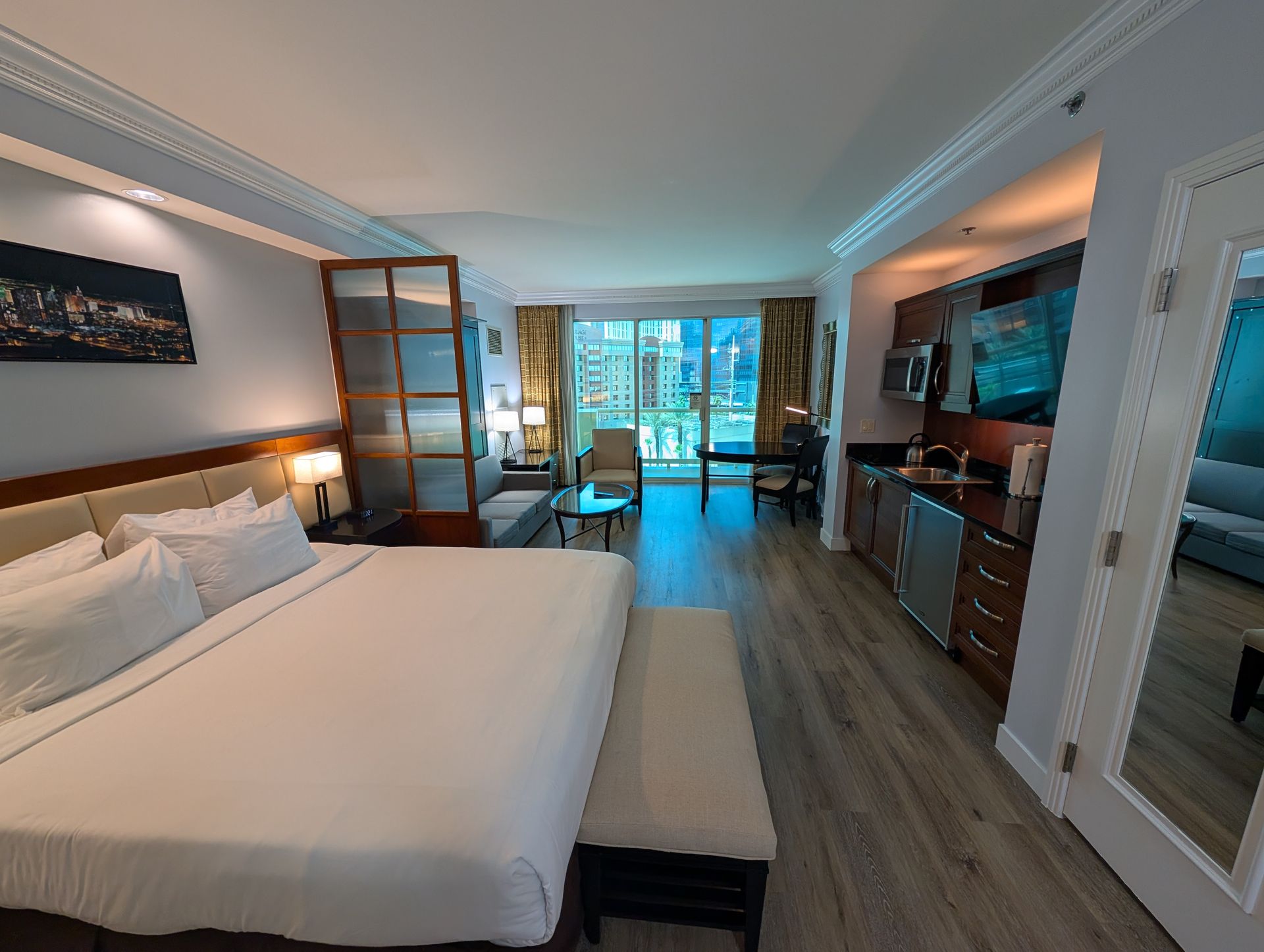 Modern hotel room with king bed, desk by balcony doors, TV, and wood furnishings