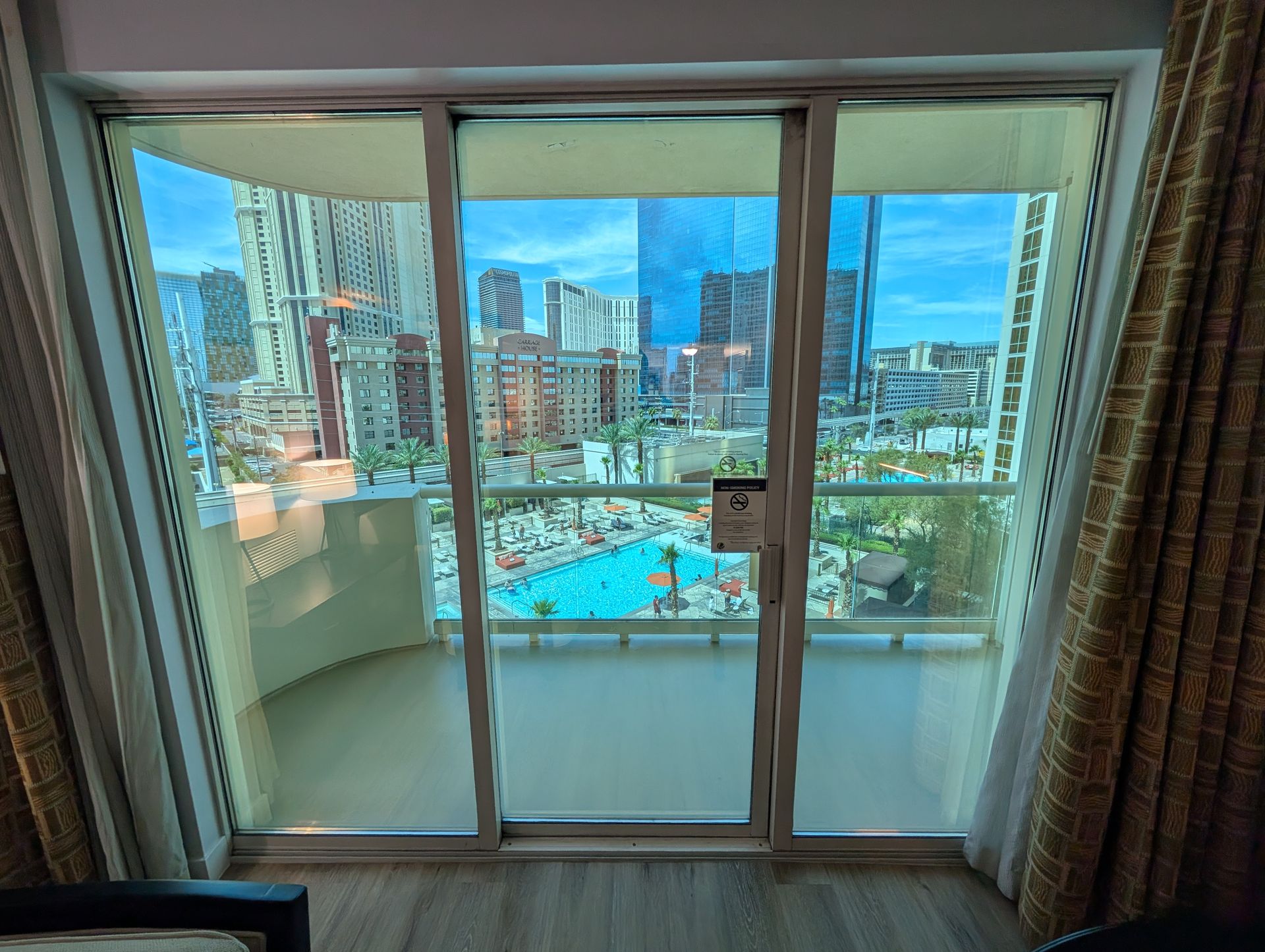 View through sliding glass doors to a city skyline and pool, framed by curtains and balcony railing