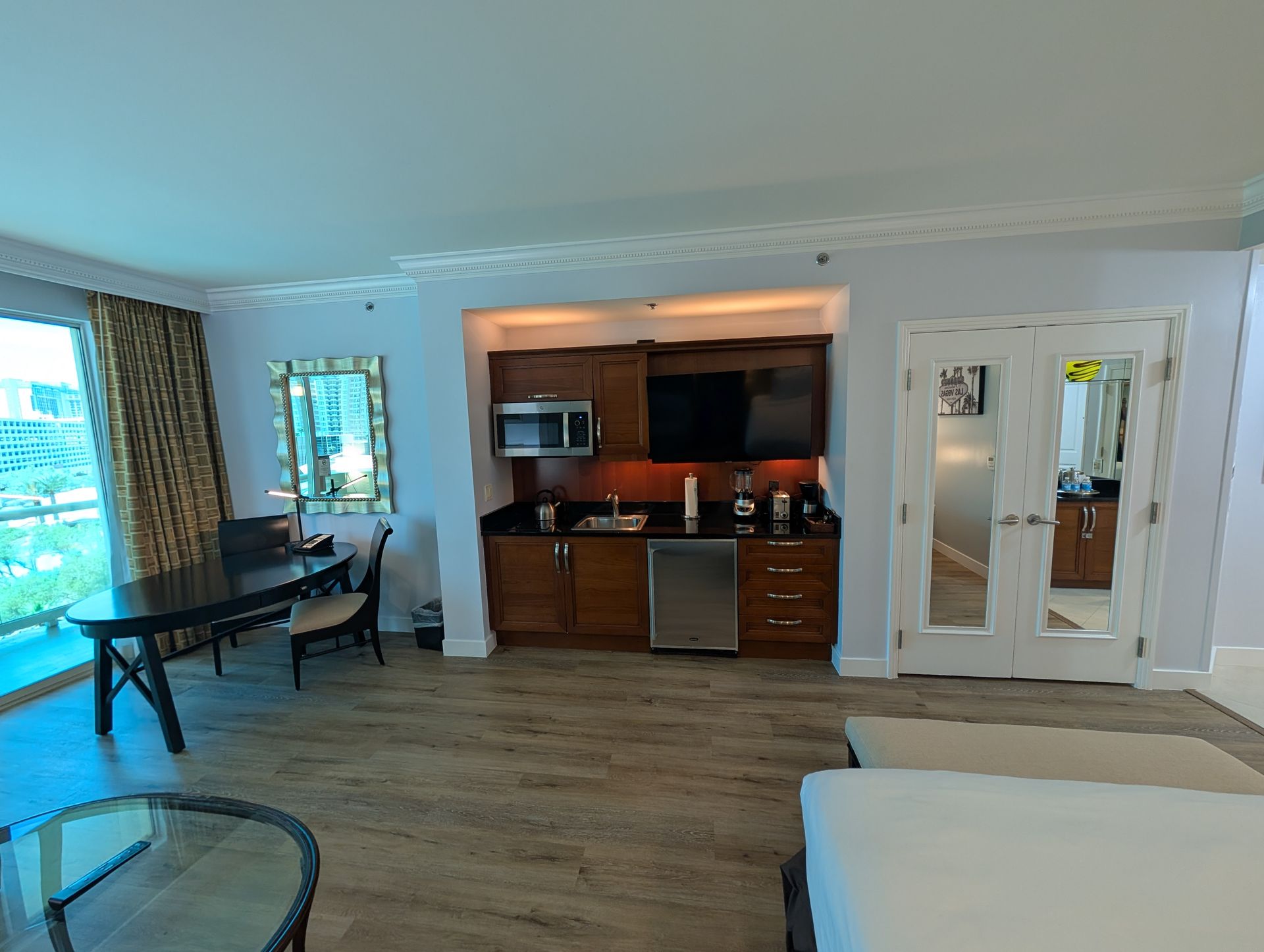 Bright hotel-style room with kitchenette, desk, sofa, and large windows overlooking greenery.
