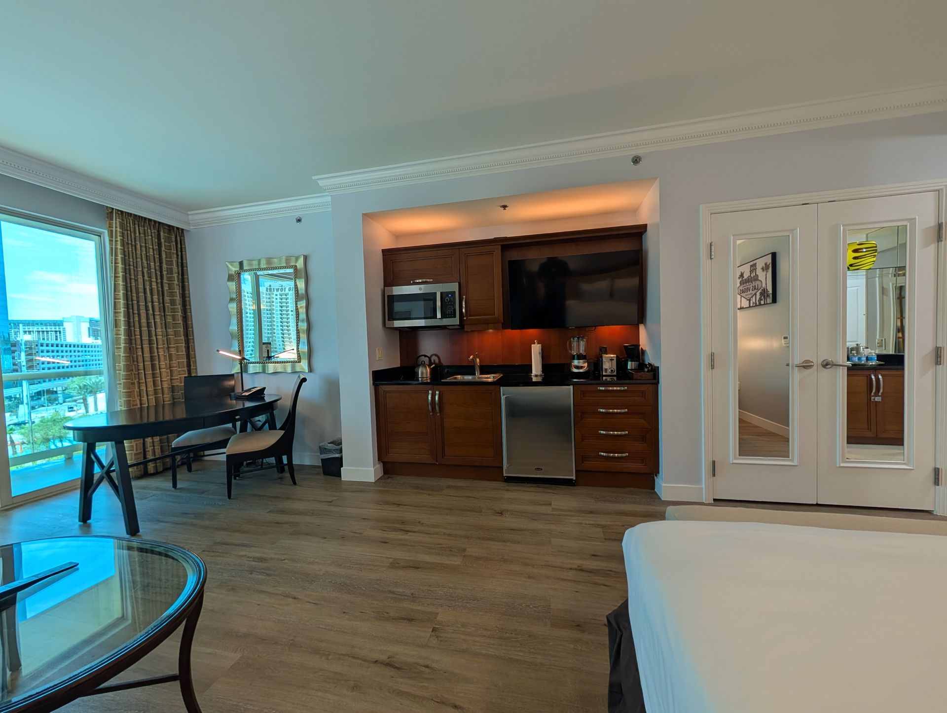 Modern hotel room with king bed, wood floors, kitchenette, dining table, and ocean view balcony