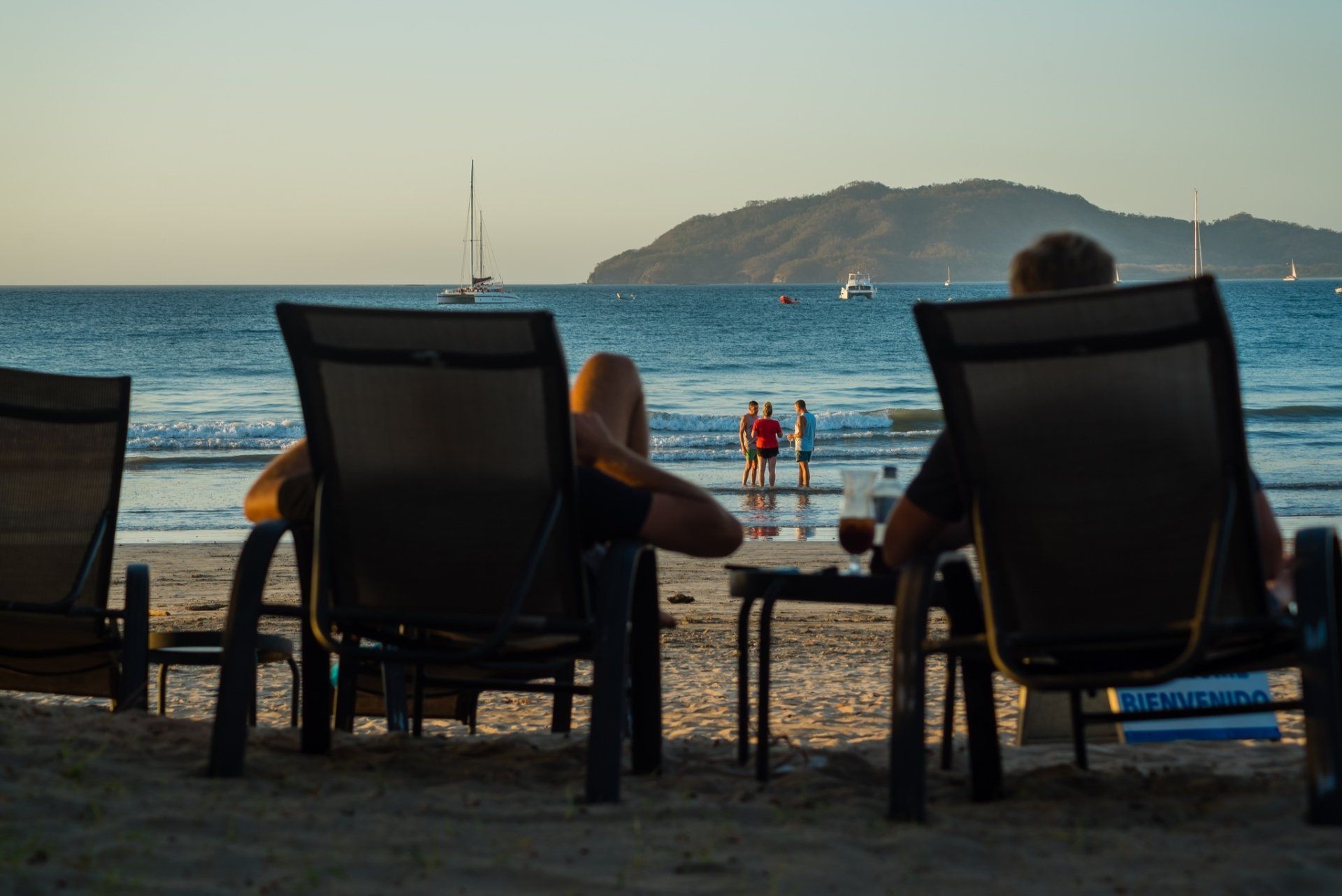 Beach lounge chairs facing the sea at sunset, with drinks on a table and an island in the distance