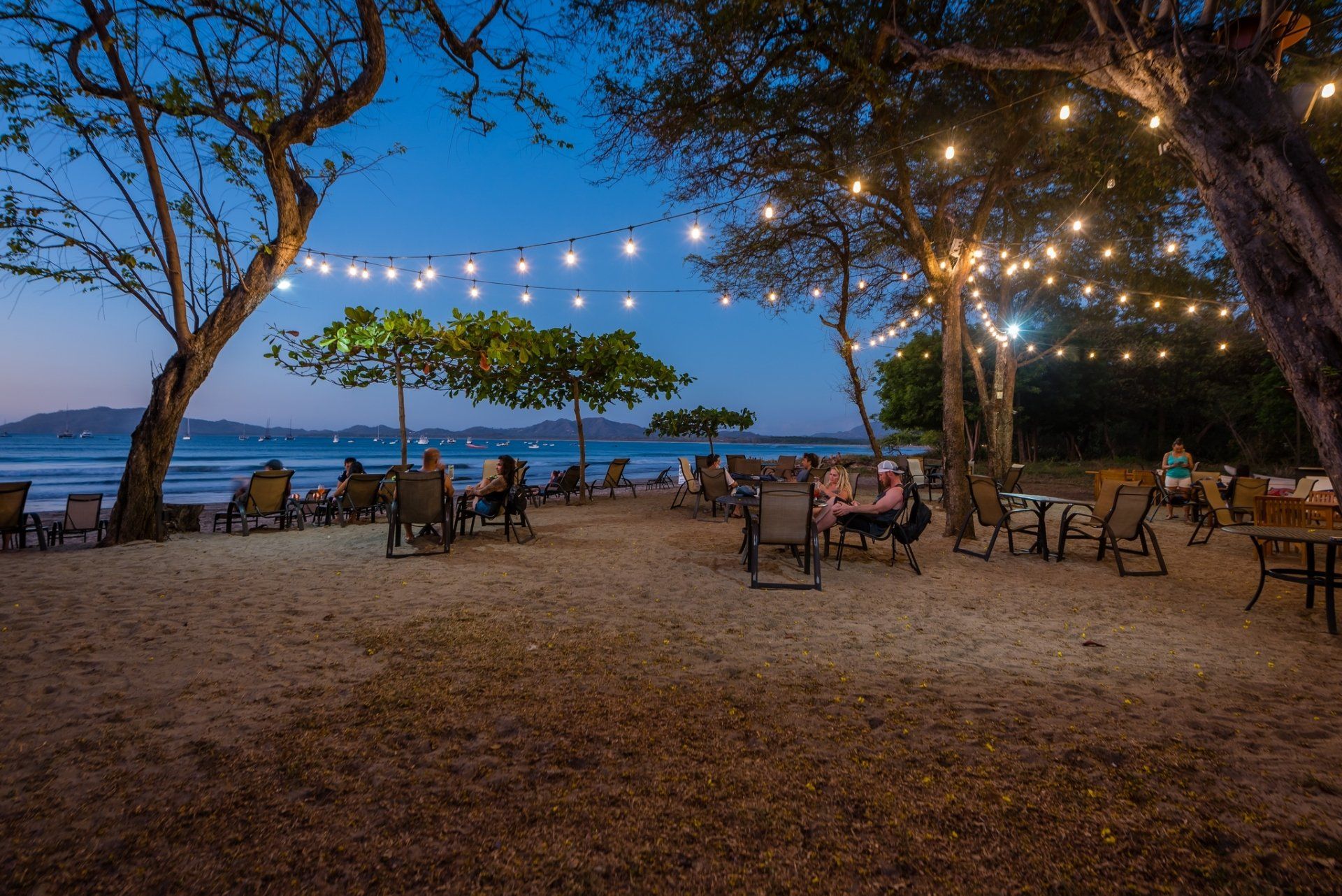 String lights over beachside dining area at dusk, with chairs and tables by the water.