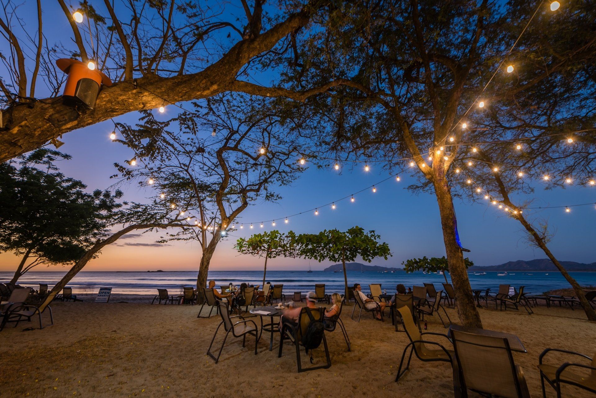 Outdoor seaside café at dusk with string lights, empty chairs, and sunset over the water