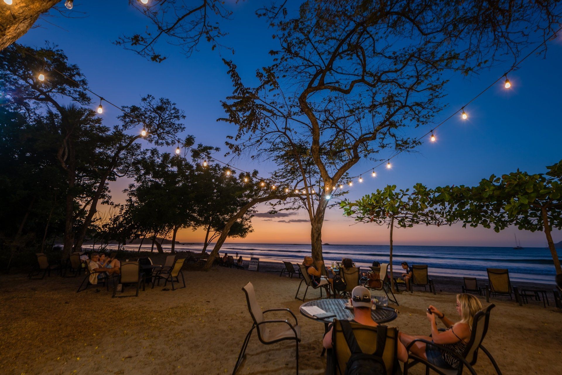 Twilight beach café with string lights, tables, and chairs overlooking the sea