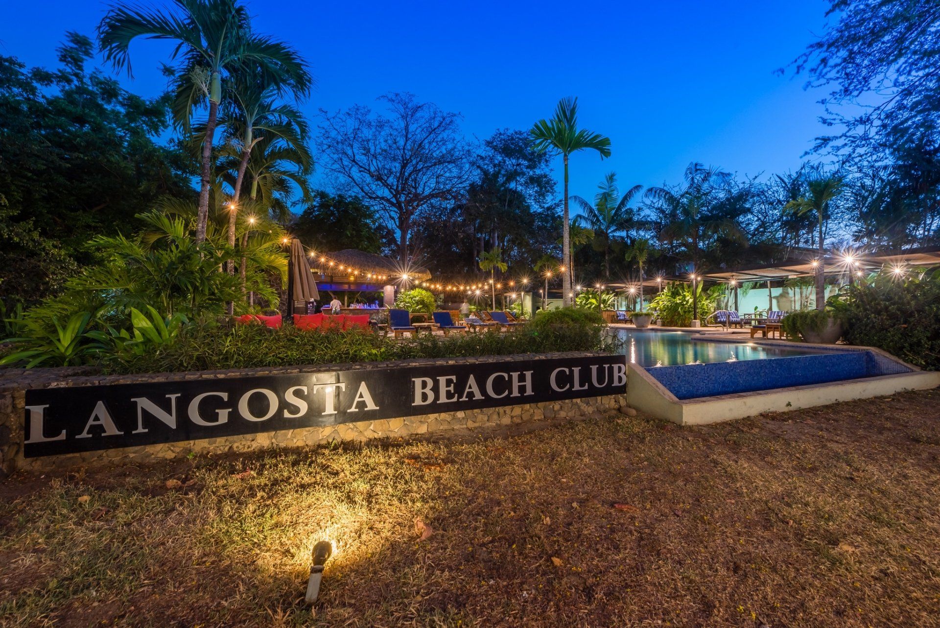 Lagosita Beach Club sign at dusk, with lit palm trees, lawn, and poolside seating in the background