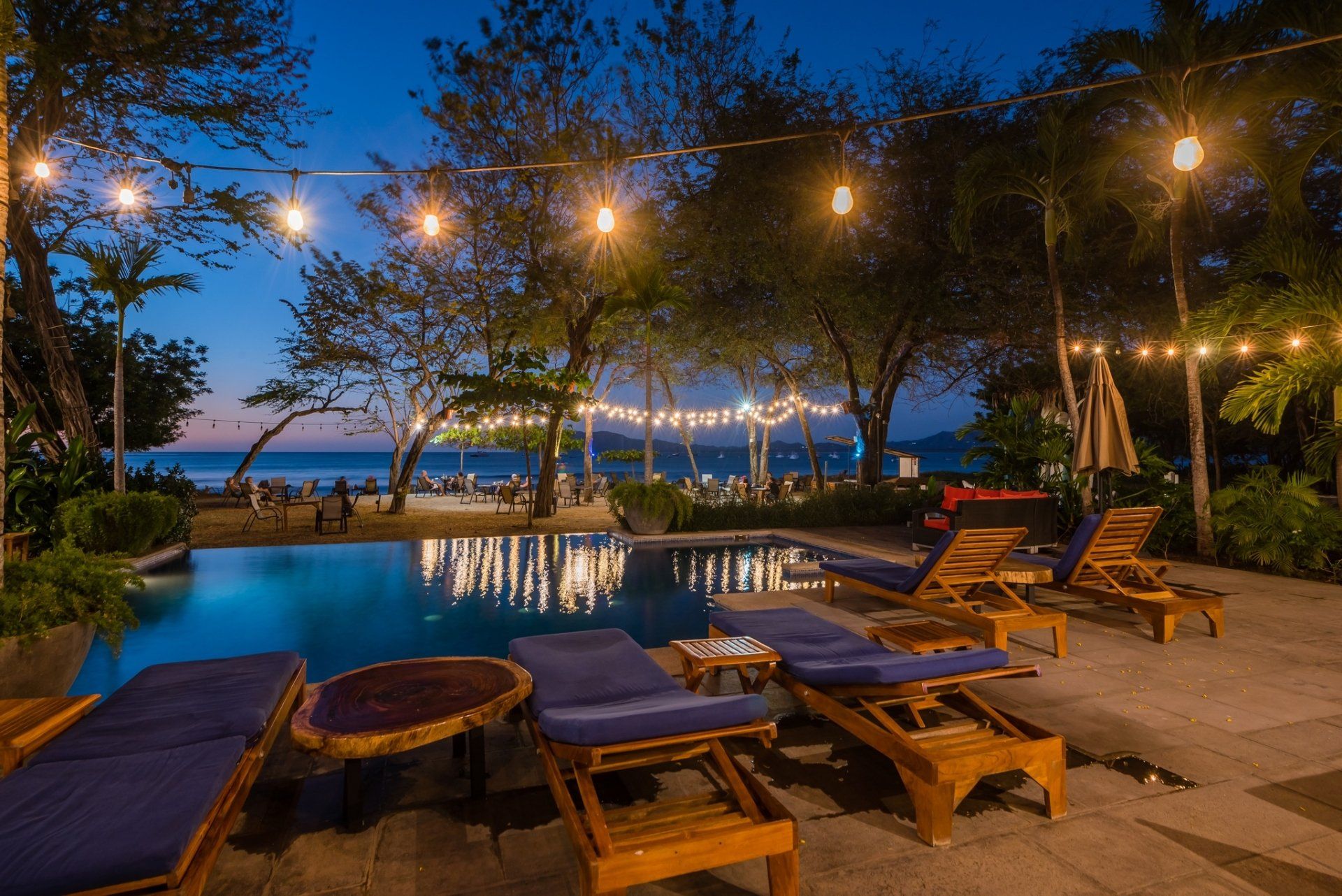 Poolside lounge with wooden chairs and string lights overlooking the ocean at dusk