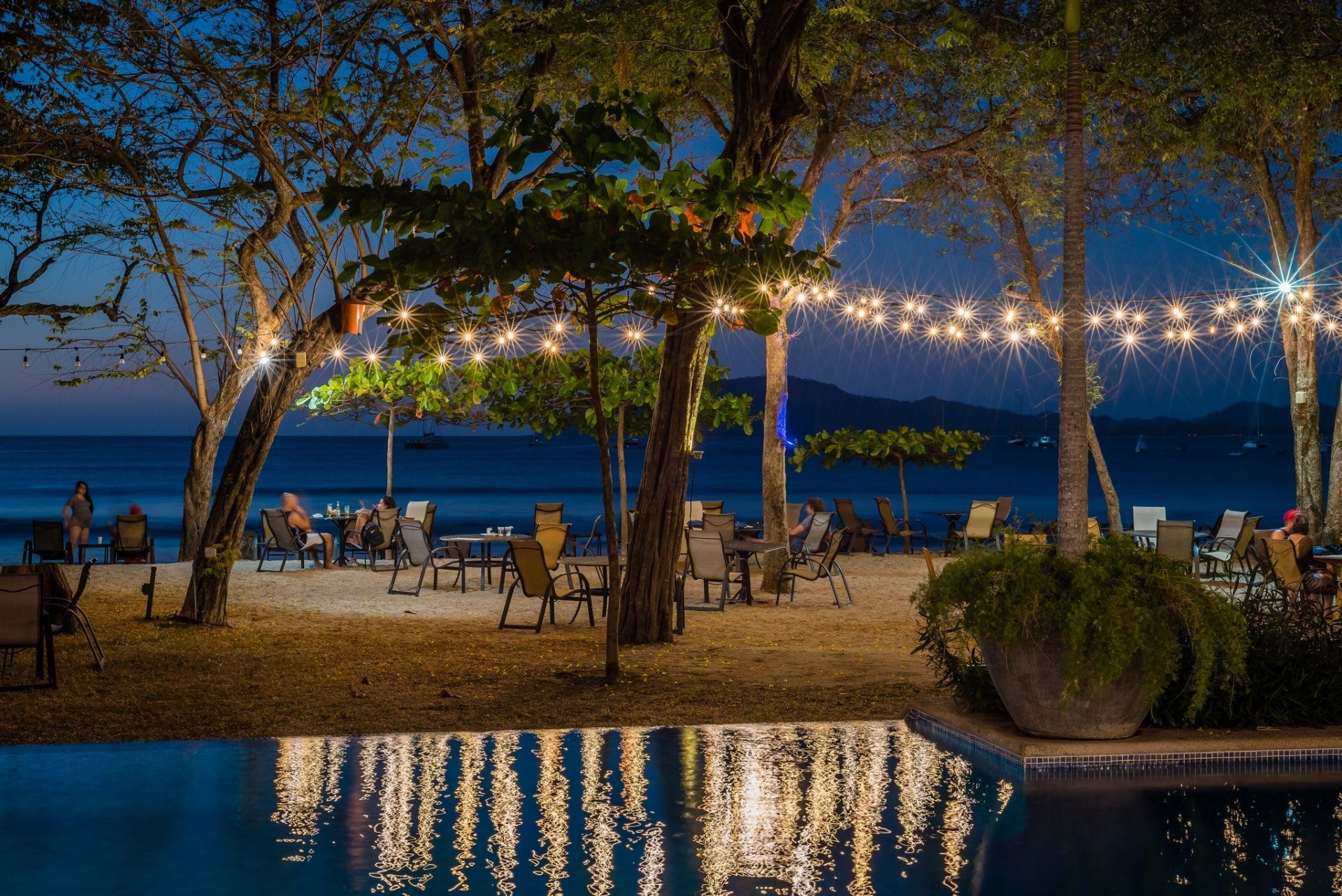 Beachfront café tables under string lights at dusk, with palm trees, ocean, and pool reflections